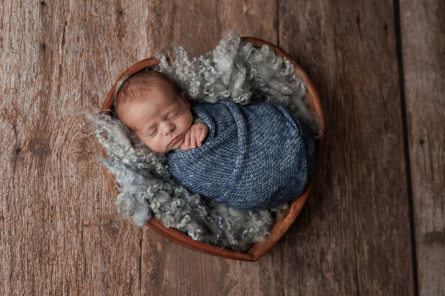 Newborn wrapped in blue cloth in a wooden bowl with soft gray fur, on a wood plank floor.
