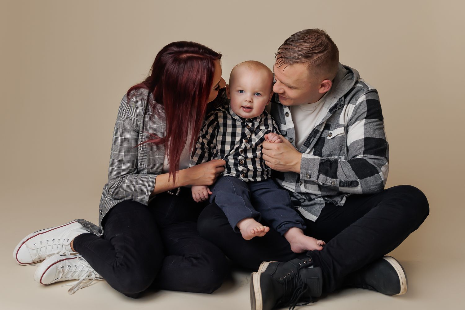 Two adults and a baby sit close together on a beige background.