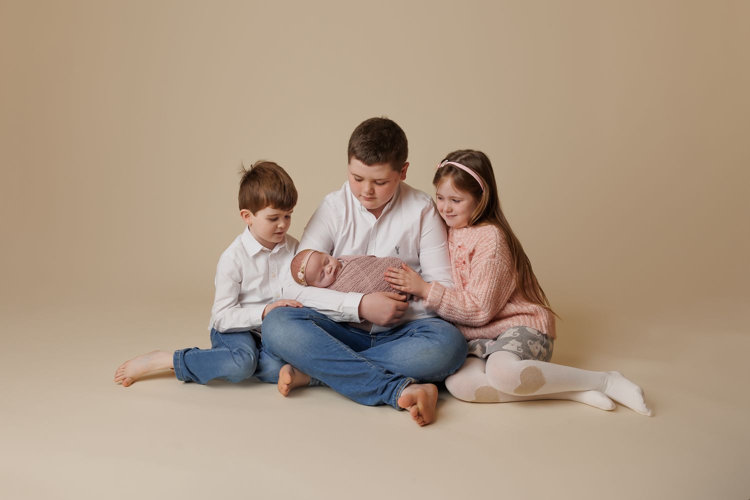 two brothers and older sister and a baby sitting together on a beige background.
