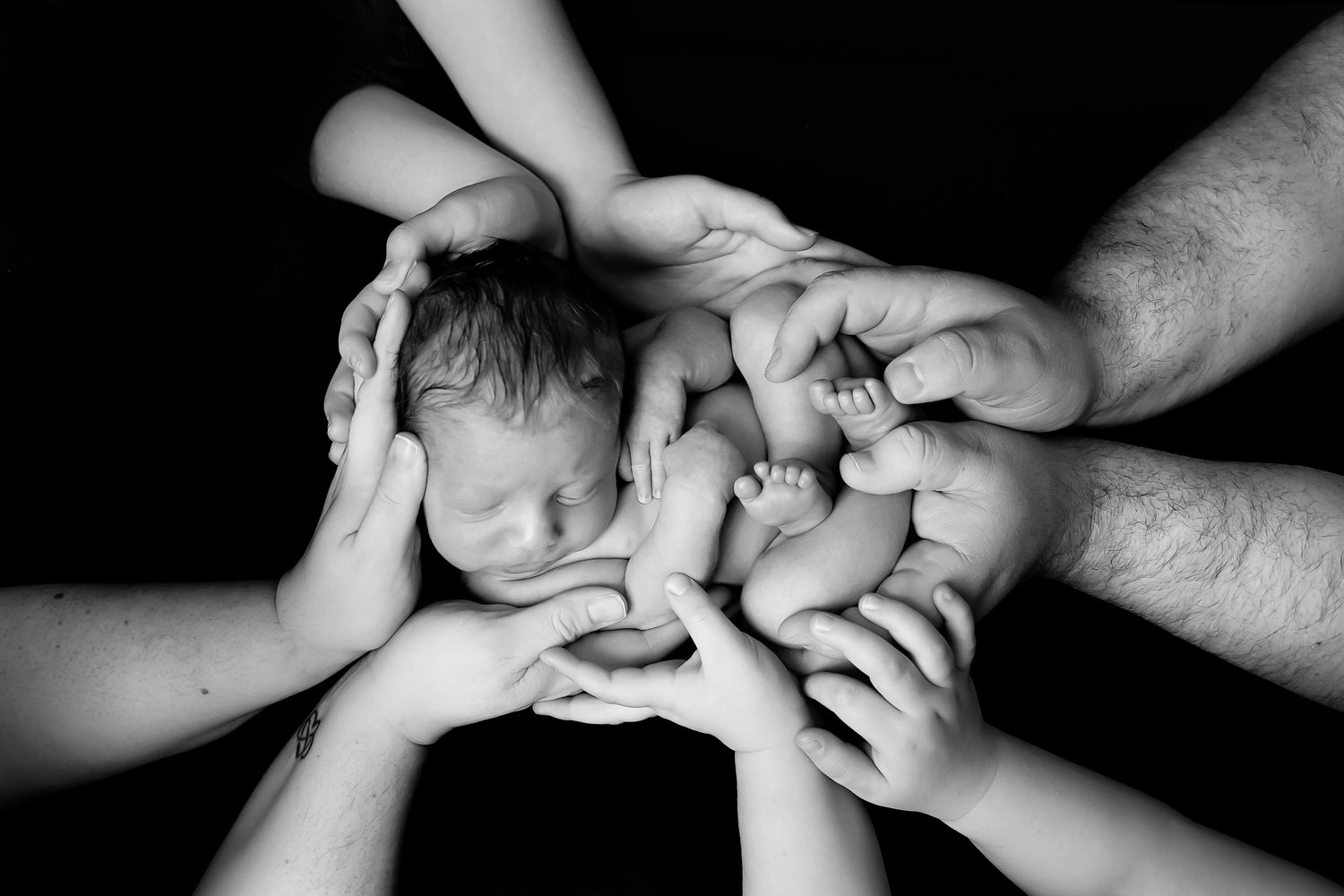 A heartwarming black and white image of a baby embraced by loving hands all around.