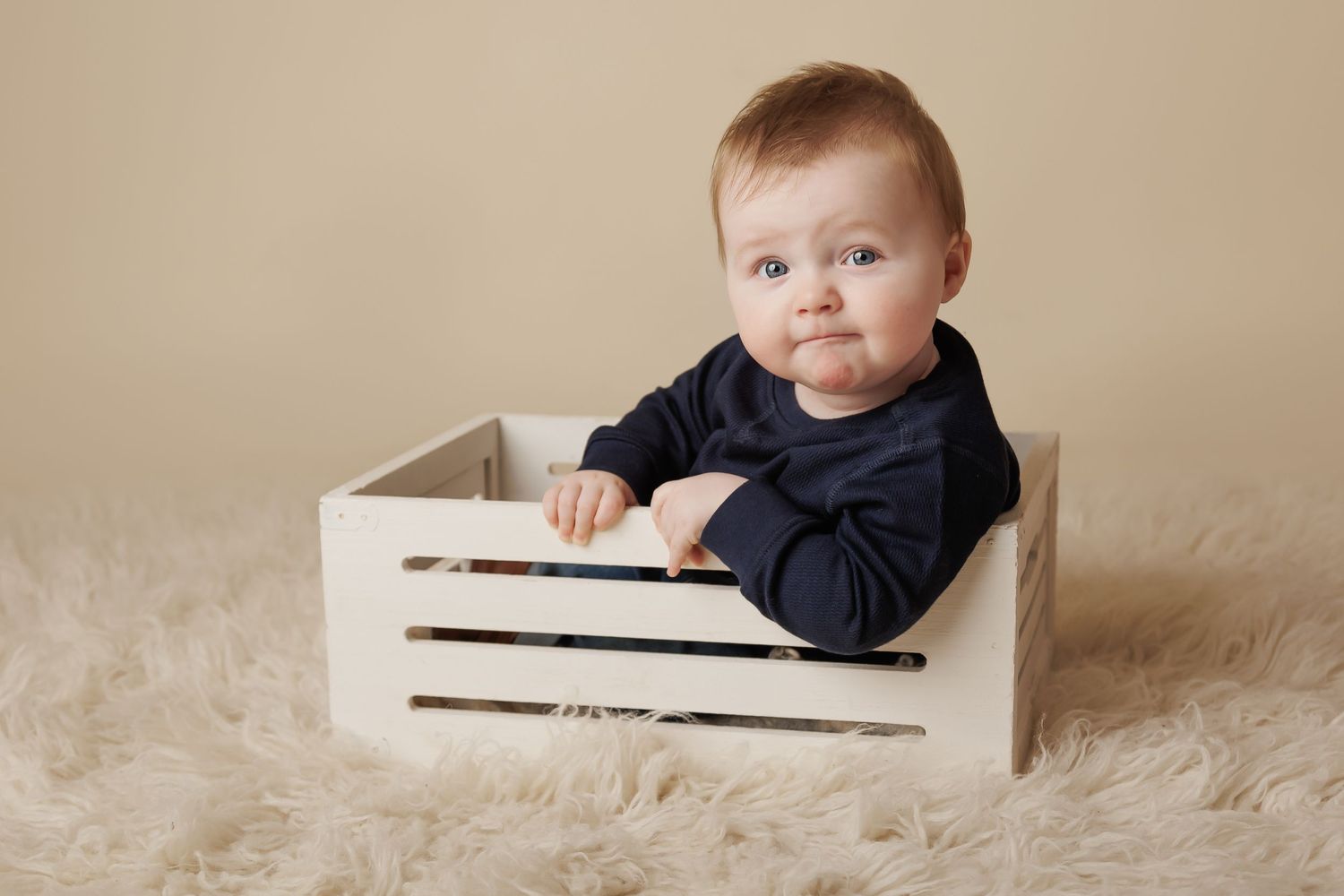 7 month old baby boy sitting in a white crate on a soft cream rug cheekily looking at the camera