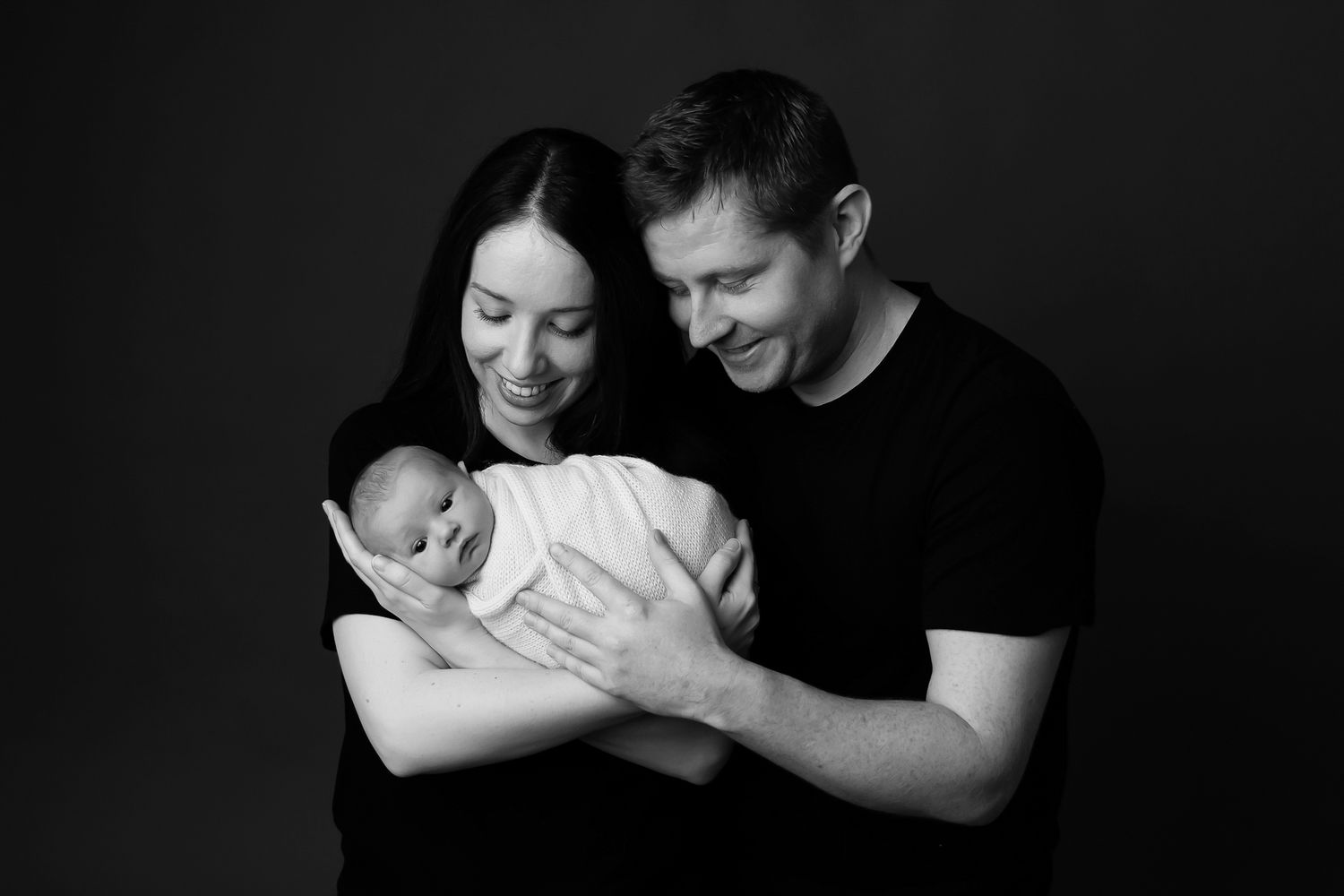 A couple smiles as they hold their baby wrapped in a blanket, standing against a plain background.
