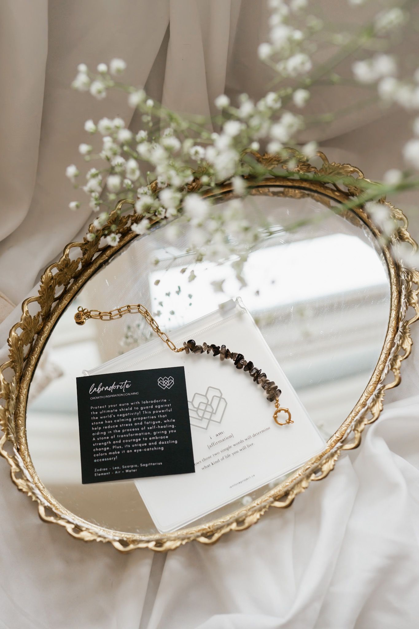 Elegant gold mirrored vanity tray with ornate edges displayed with small white flowers and product cards in soft lighting.