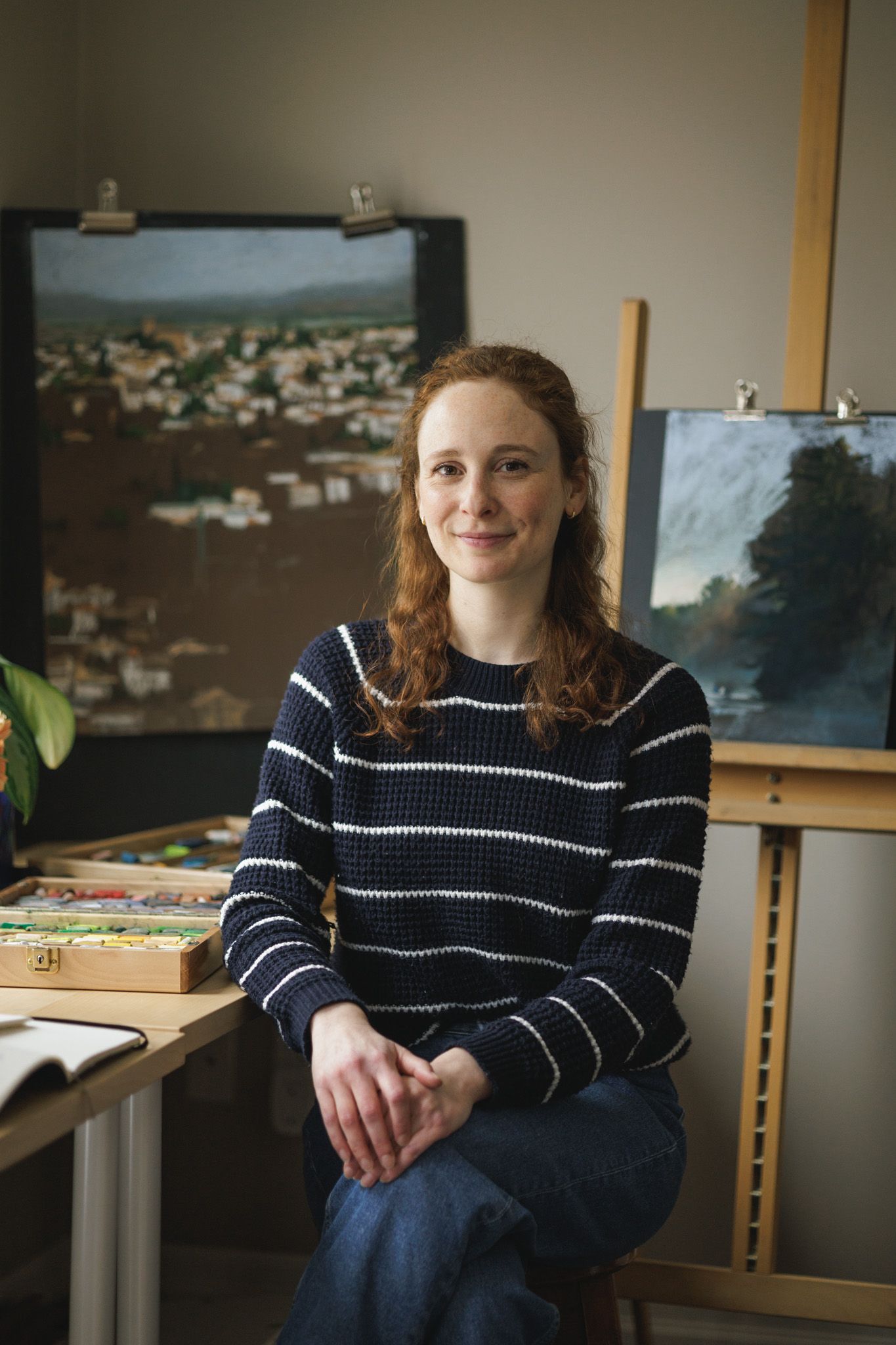 Branding portrait of an artist sitting in front of her artwork and paintings with a box of soft pastels.
