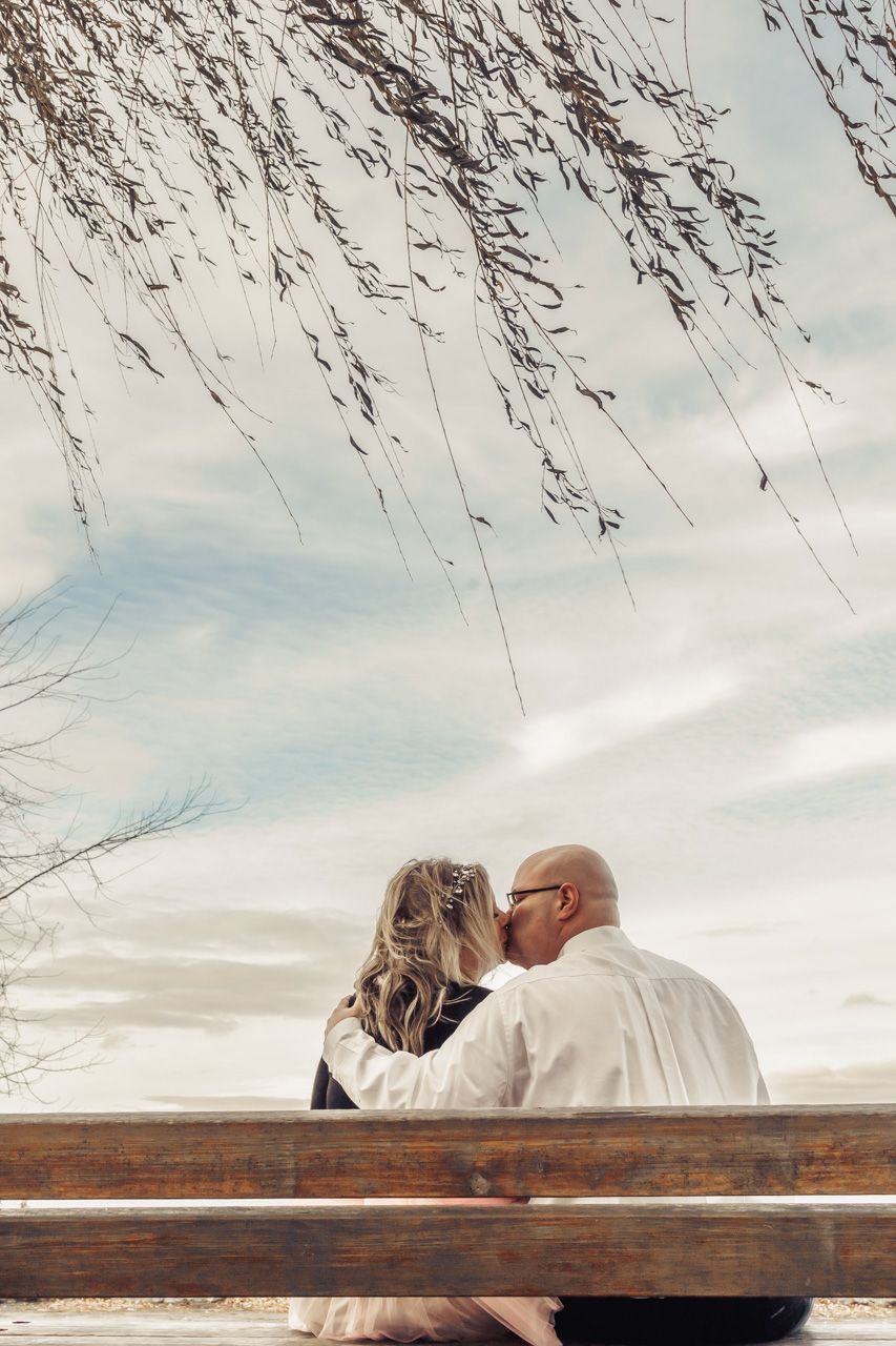 Romantic kiss between a couple captured on a wooden bench against a cloudy sky with hanging branches.
