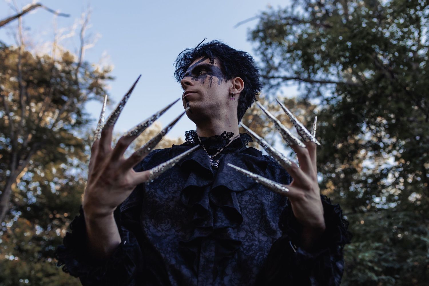 A dramatic low-angle shot of someone in dark clothing making hand gestures against tree branches.