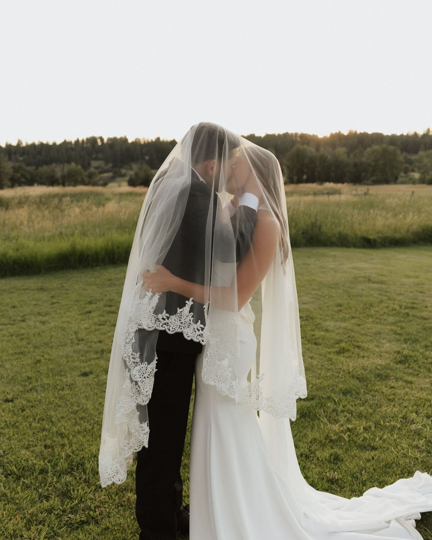 A couple shares an intimate embrace under a lace-trimmed veil in a rustic outdoor setting.