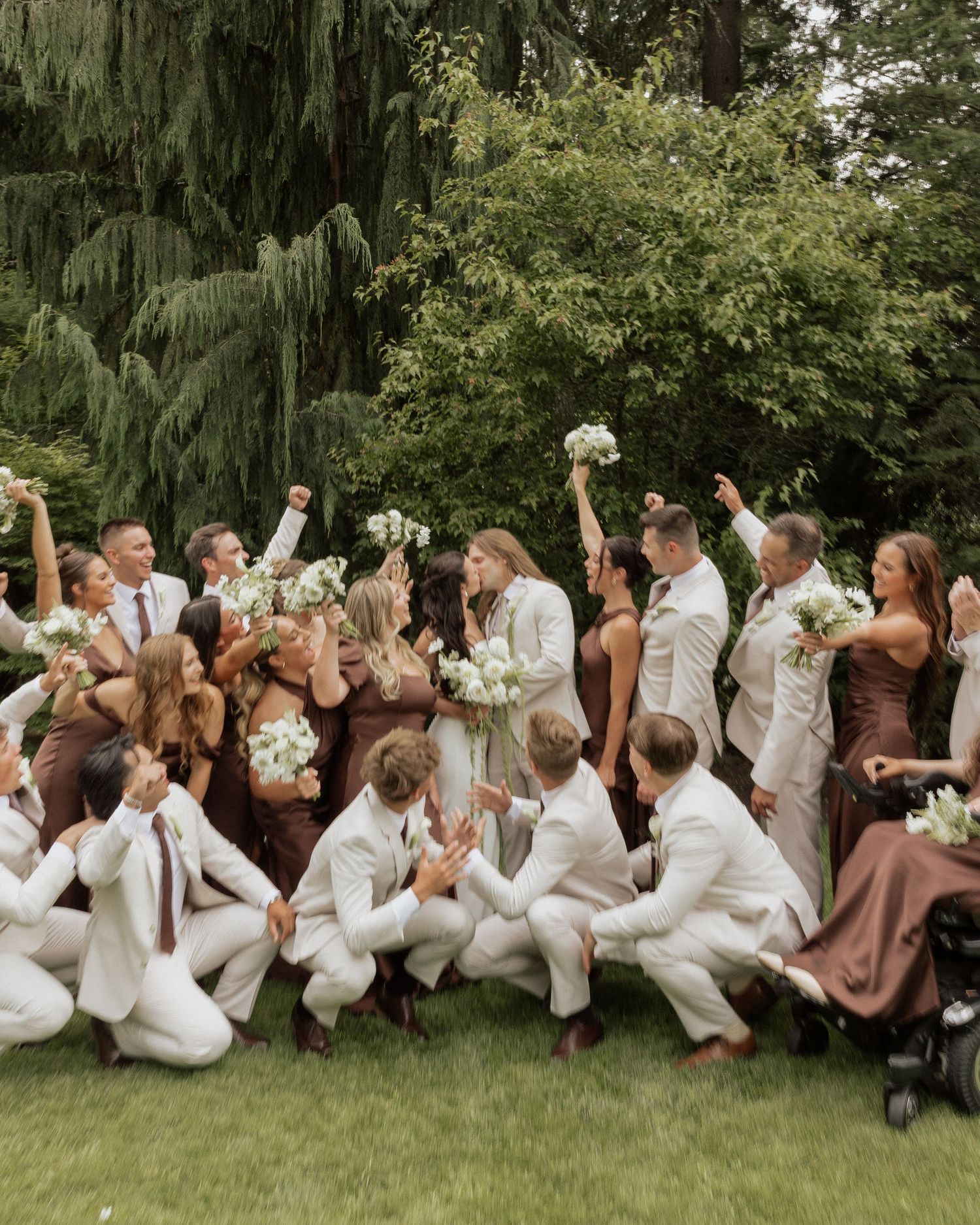 Large wedding party in cream and brown attire celebrate outdoors with bouquets.