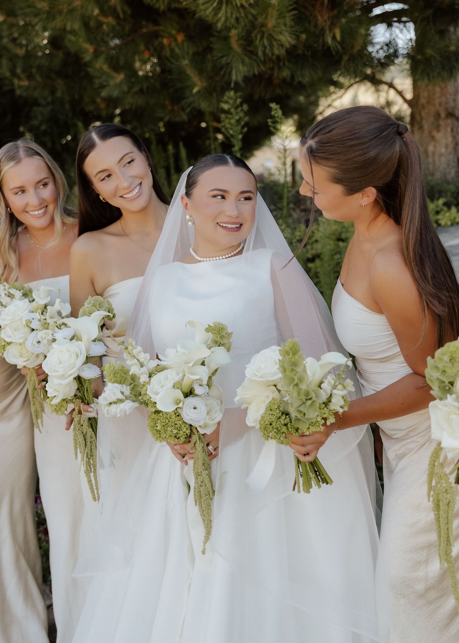 Bride in white gown and veil poses with bridesmaids in champagne dresses holding white and green bouquets.