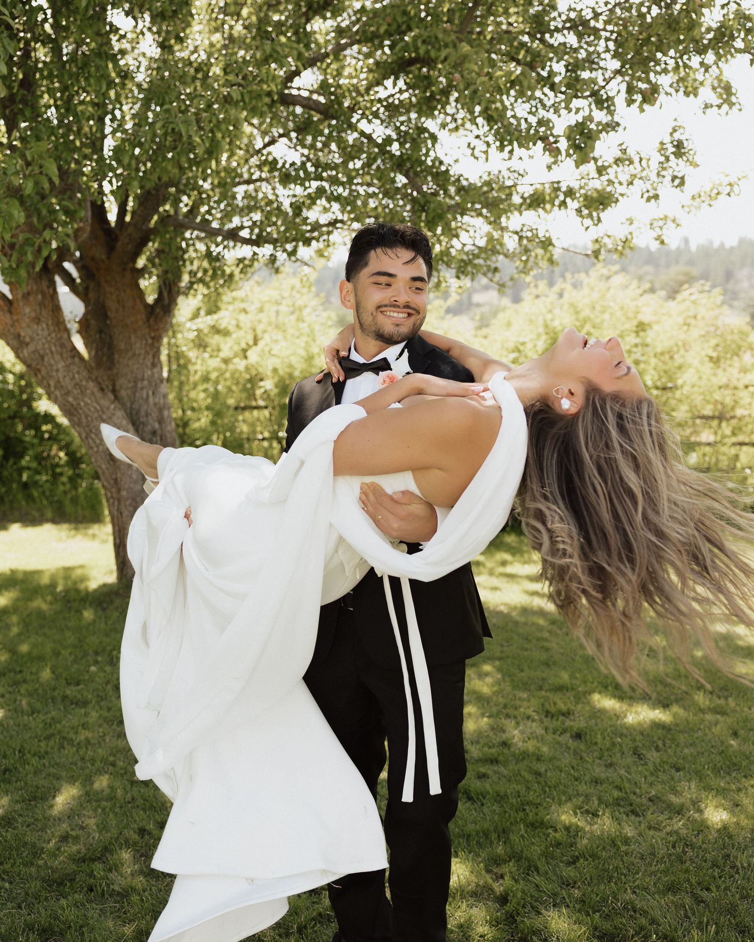 Groom in black tuxedo lifts bride in white gown outdoors under green trees at wedding celebration.