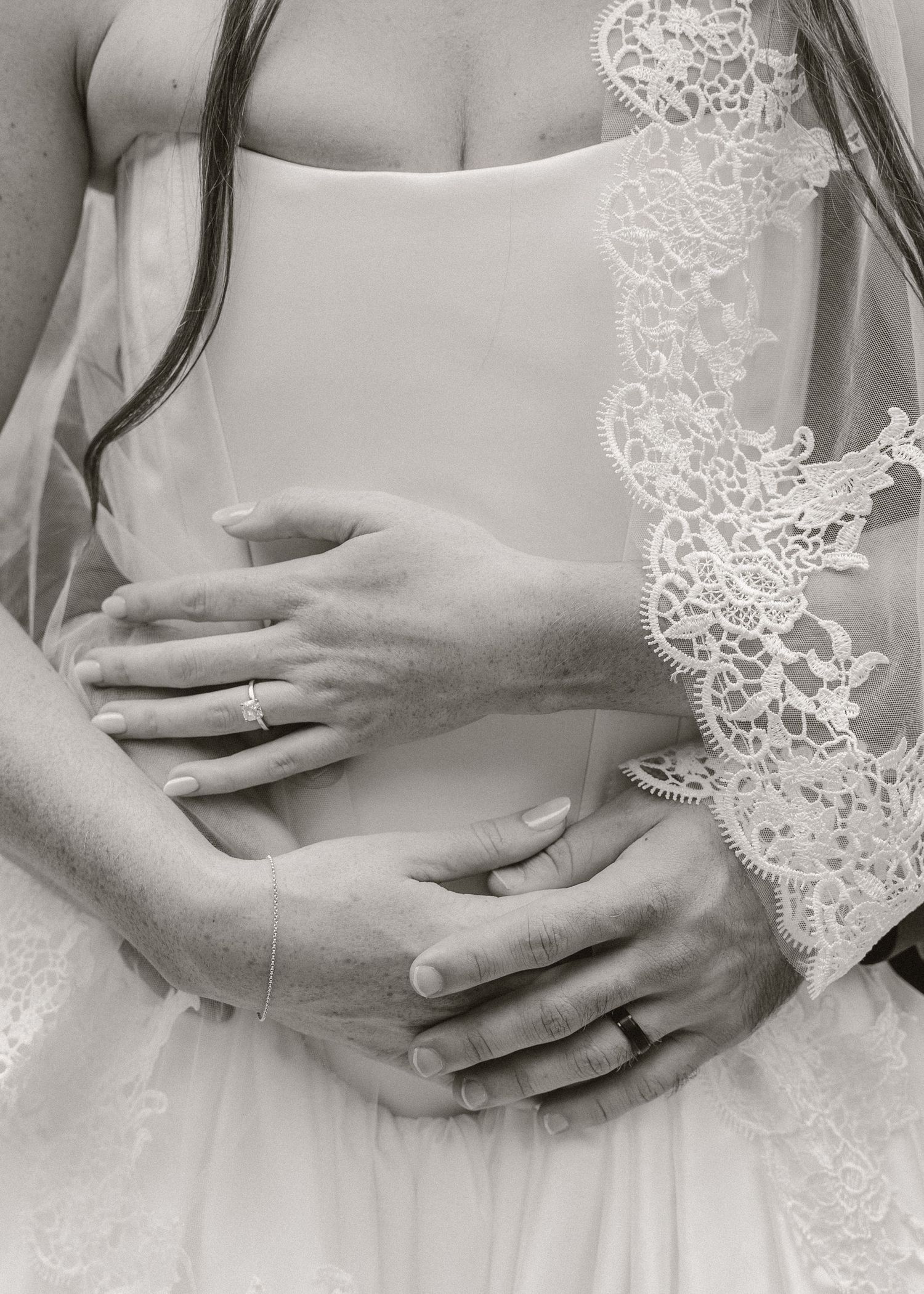 Black and white close-up of bride and groom hands embracing, featuring engagement ring and lace wedding veil detail.