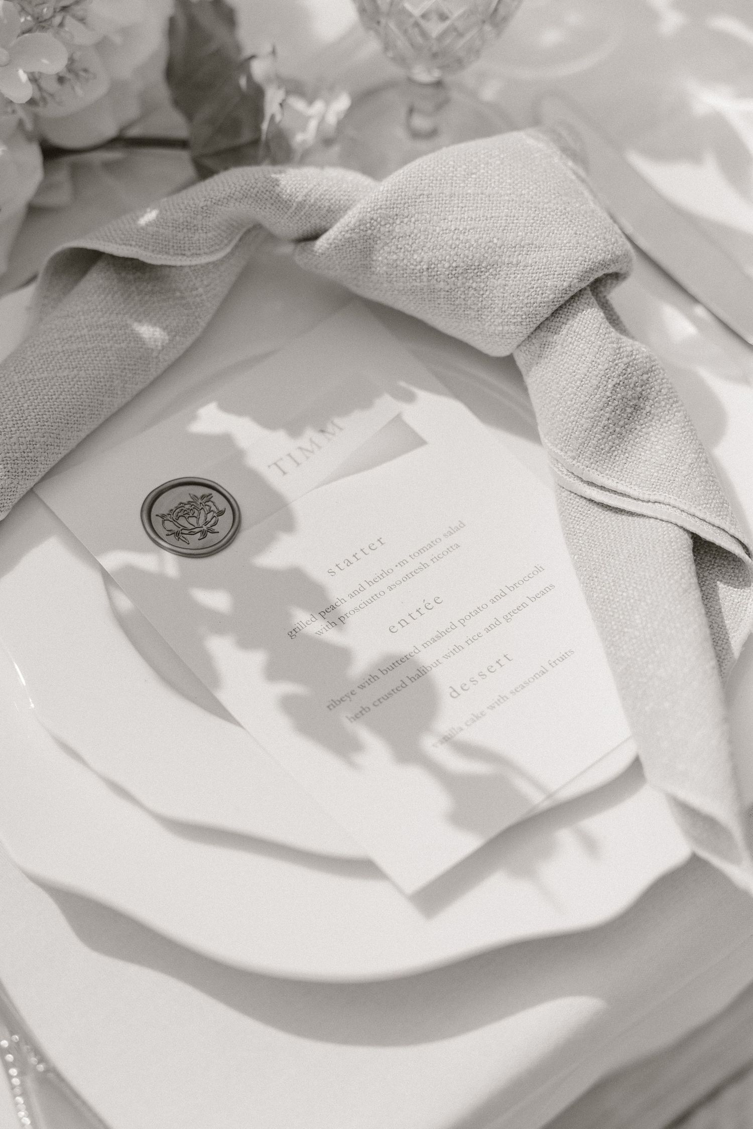 Elegant wedding place setting with white plates, gray linen napkin, and wax-sealed menu card in soft natural light.