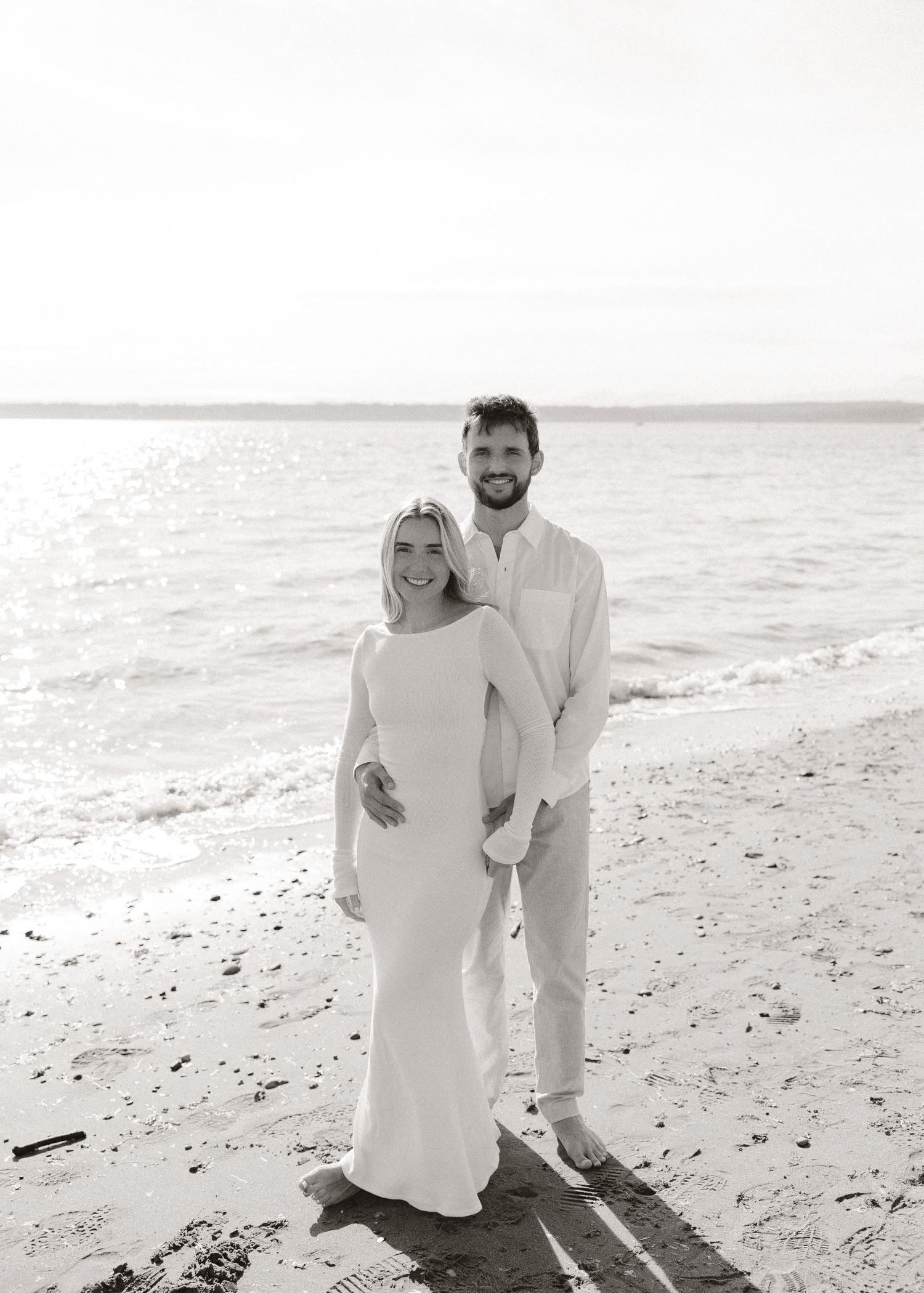 Couple in white attire posing together on a sandy beach with ocean waves in the background.