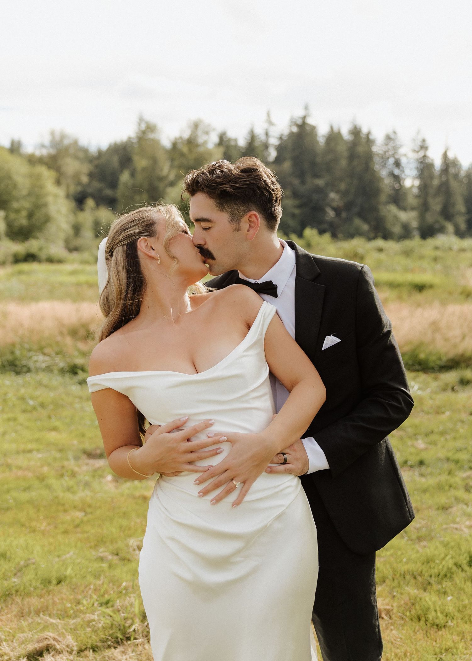 Bride and groom sharing a romantic kiss outdoors in a green meadow, wearing elegant white gown and black tuxedo.
