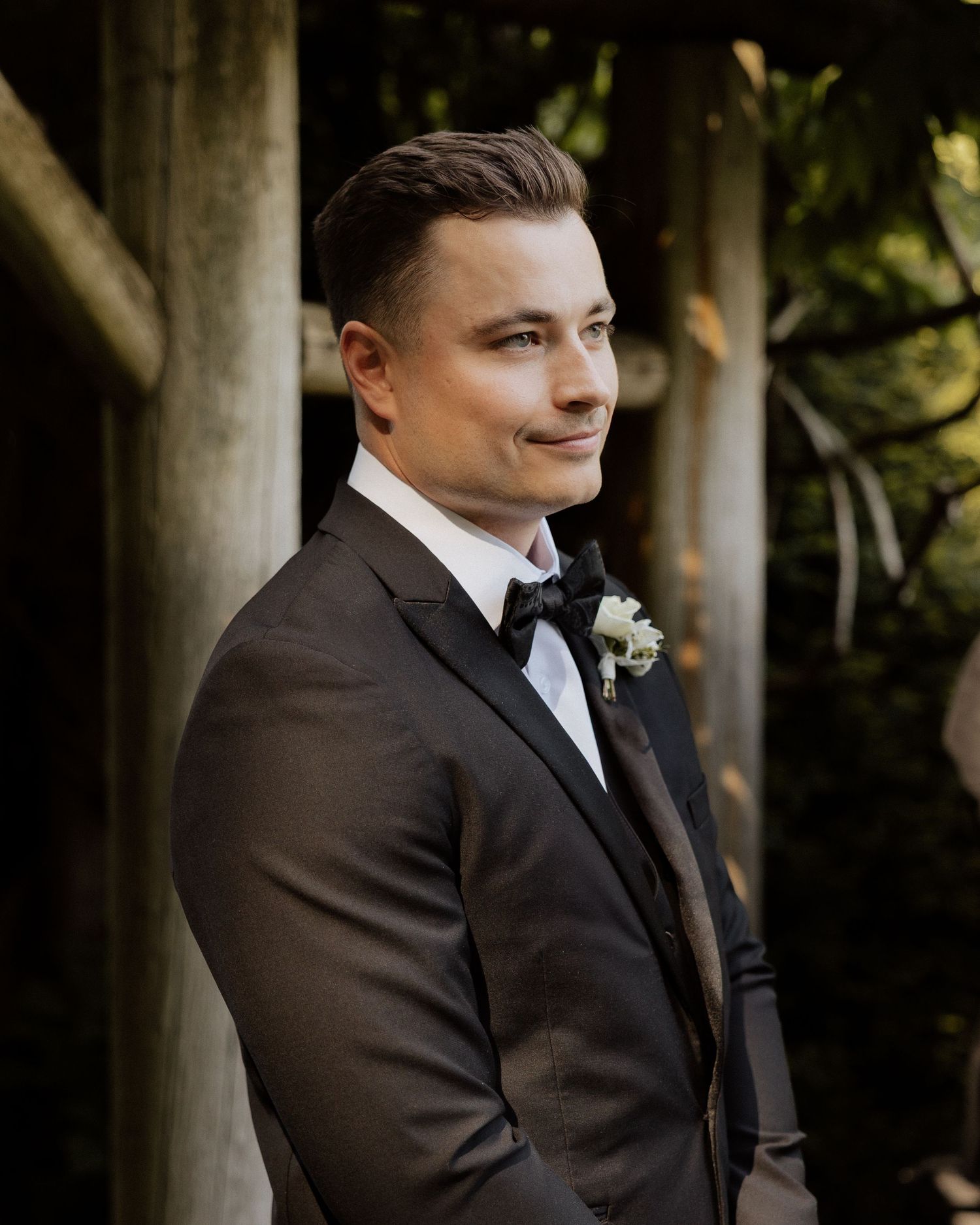 Handsome groom in dark tuxedo with white boutonniere smiling softly at an outdoor wedding venue.