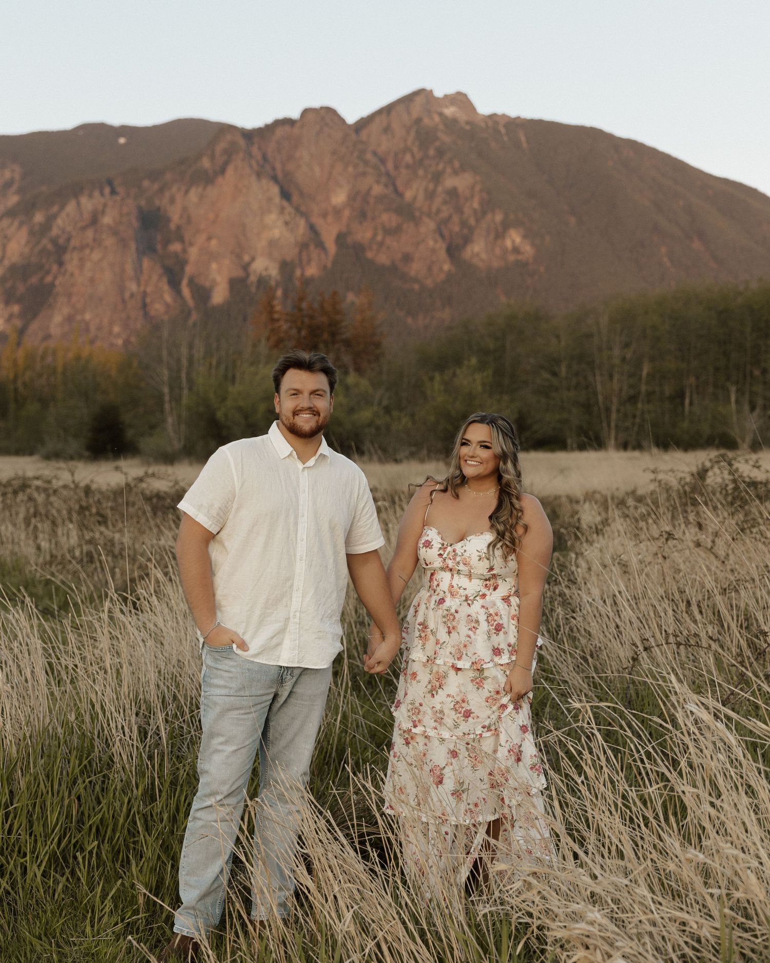 Couple holds hands while standing in tall grass with mountain scenery.