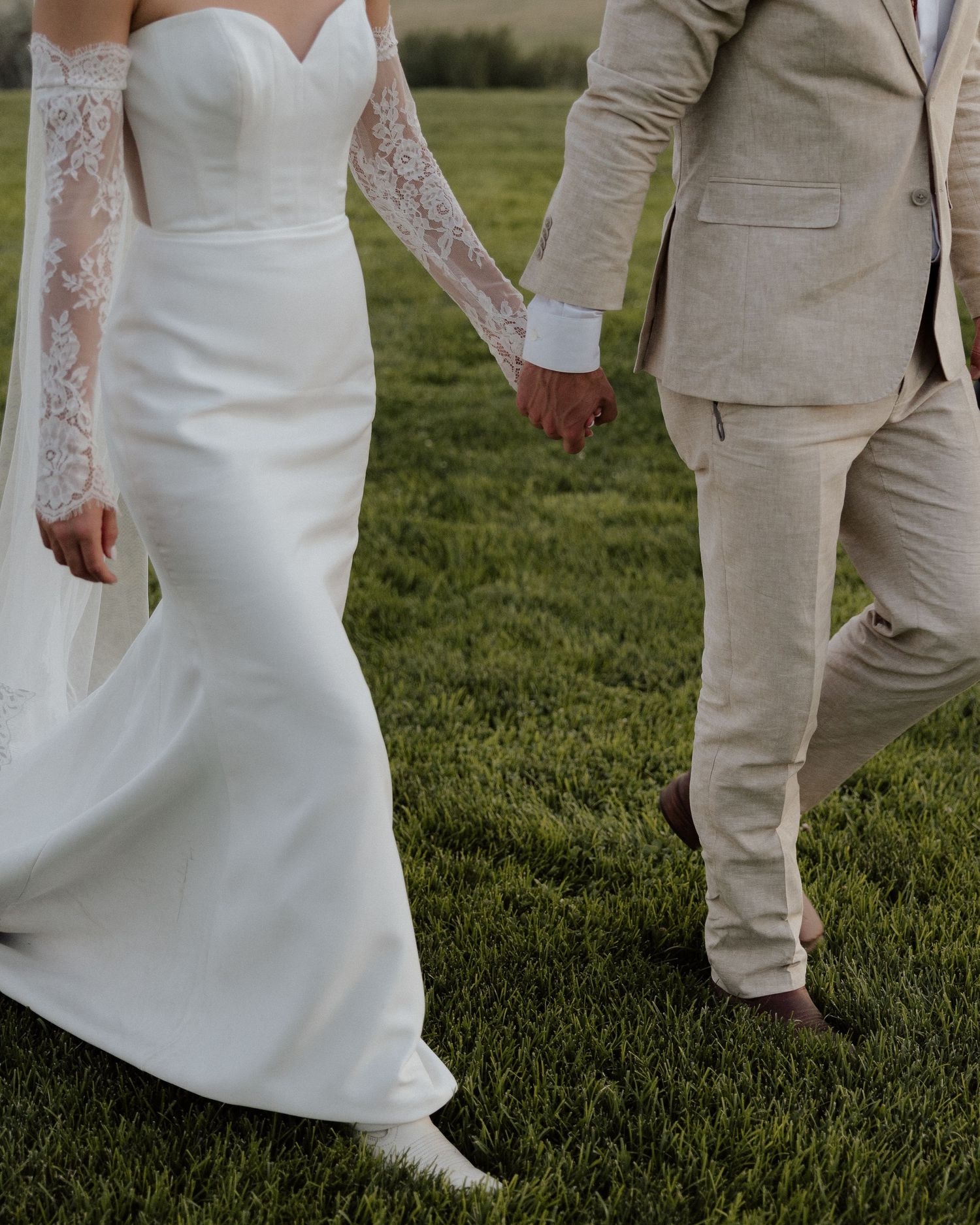 Detail shot of bride in lace sleeve gown walking beside groom in beige suit on grassy hillside.