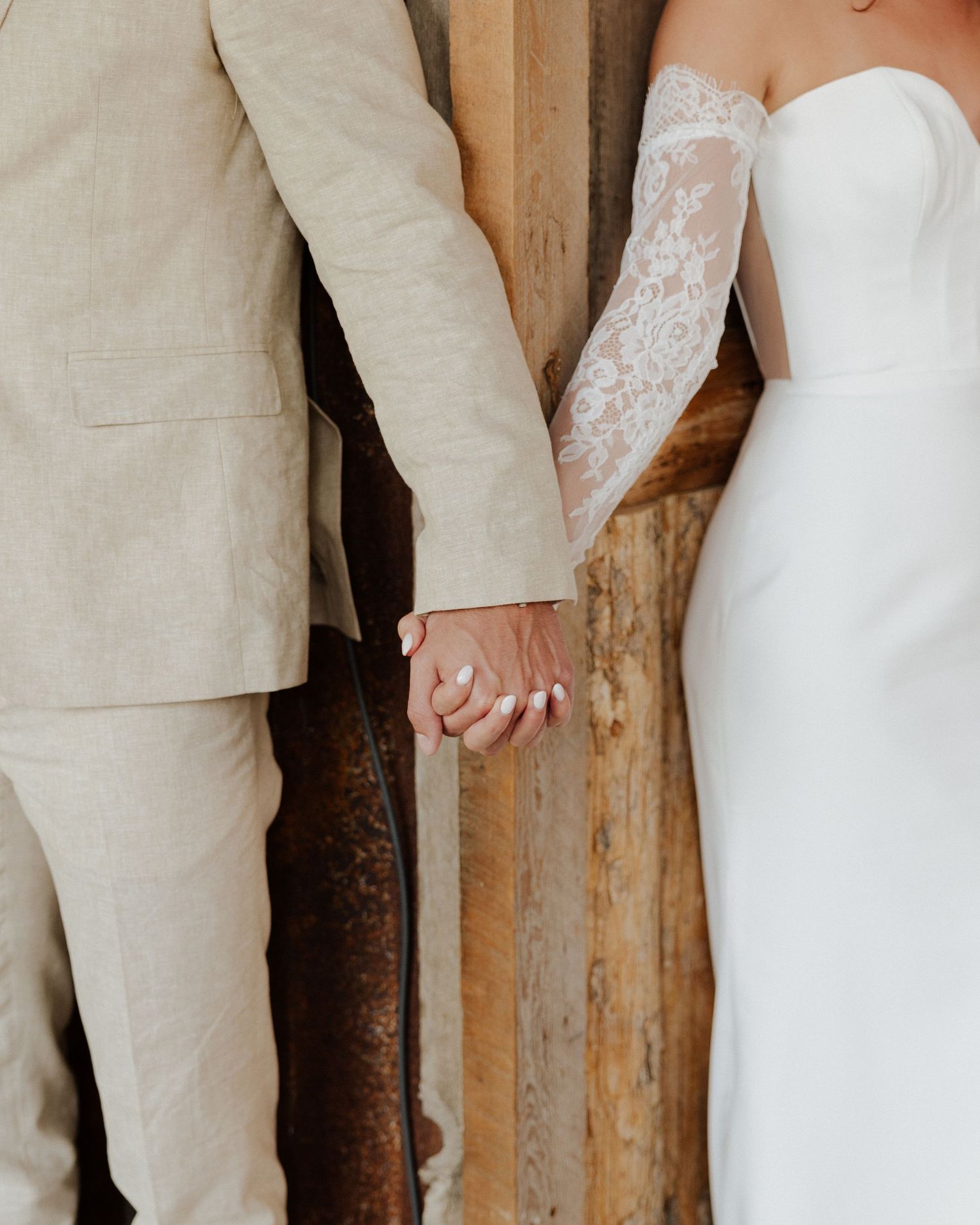 Close-up of bride and groom holding hands, showing white lace gown and beige suit details.