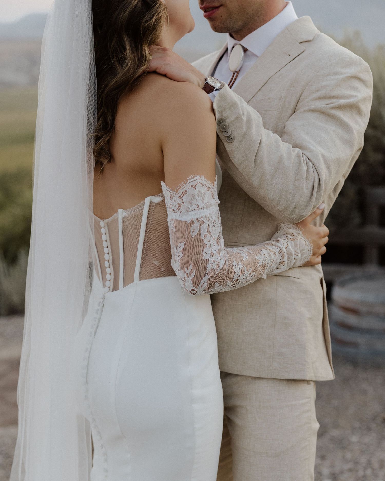 Close-up shot of newlyweds embracing, bride wearing a detailed lace dress and groom in beige suit.