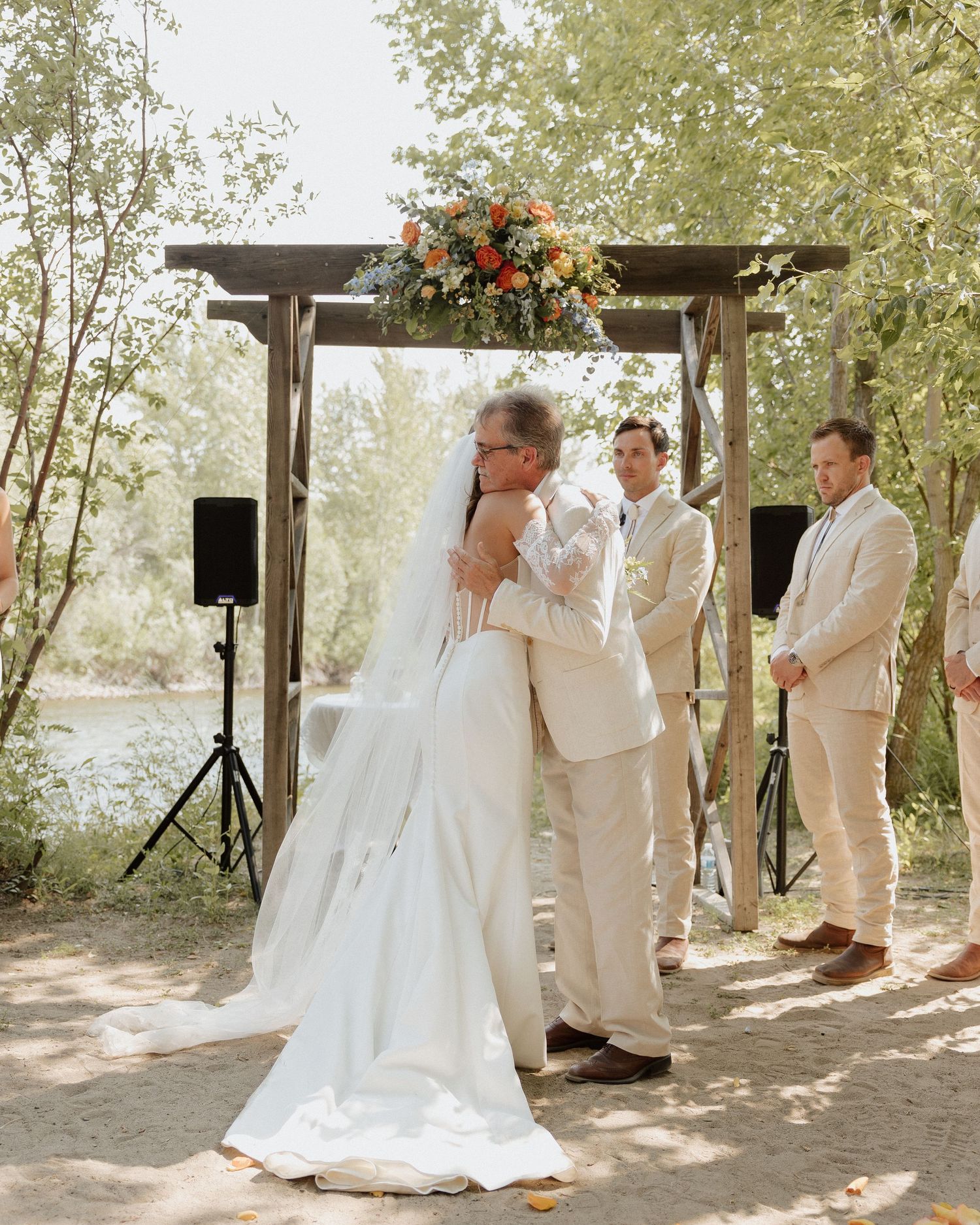 Wedding ceremony under wooden arch decorated with colorful floral arrangement and surrounded by trees.