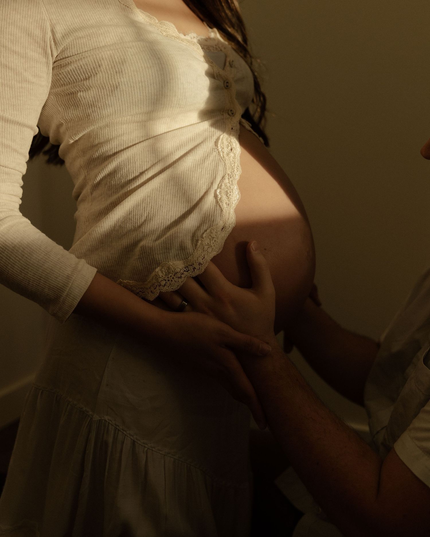 Side view of a pregnant woman in a white lace dress holding her belly in soft lighting.