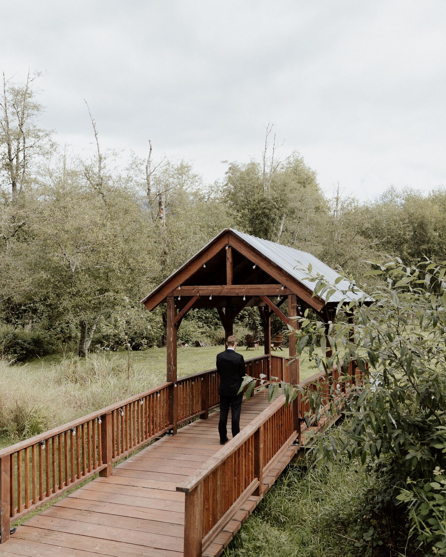 Rustic covered wooden bridge pathway surrounded by lush green trees on an overcast day.