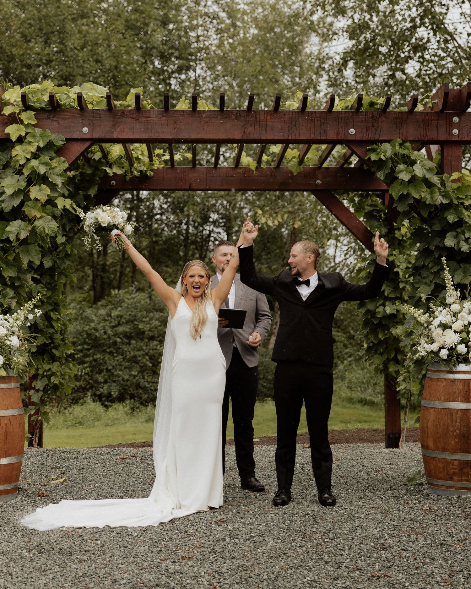 Newlyweds celebrate with raised arms under a garden arch with wine barrels.