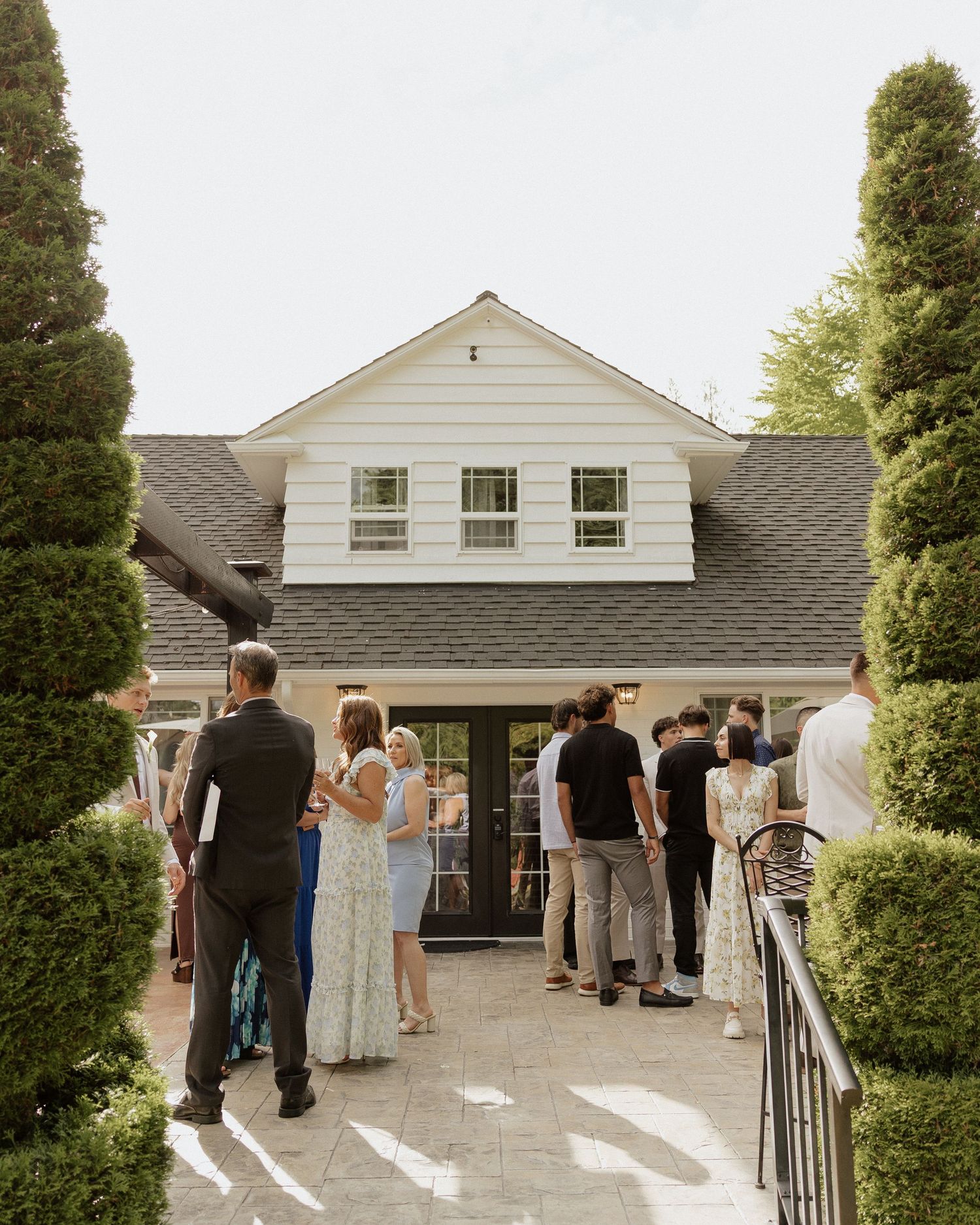 Guests gather outside a white clapboard house with dark shutters, surrounded by manicured hedges at an outdoor event.