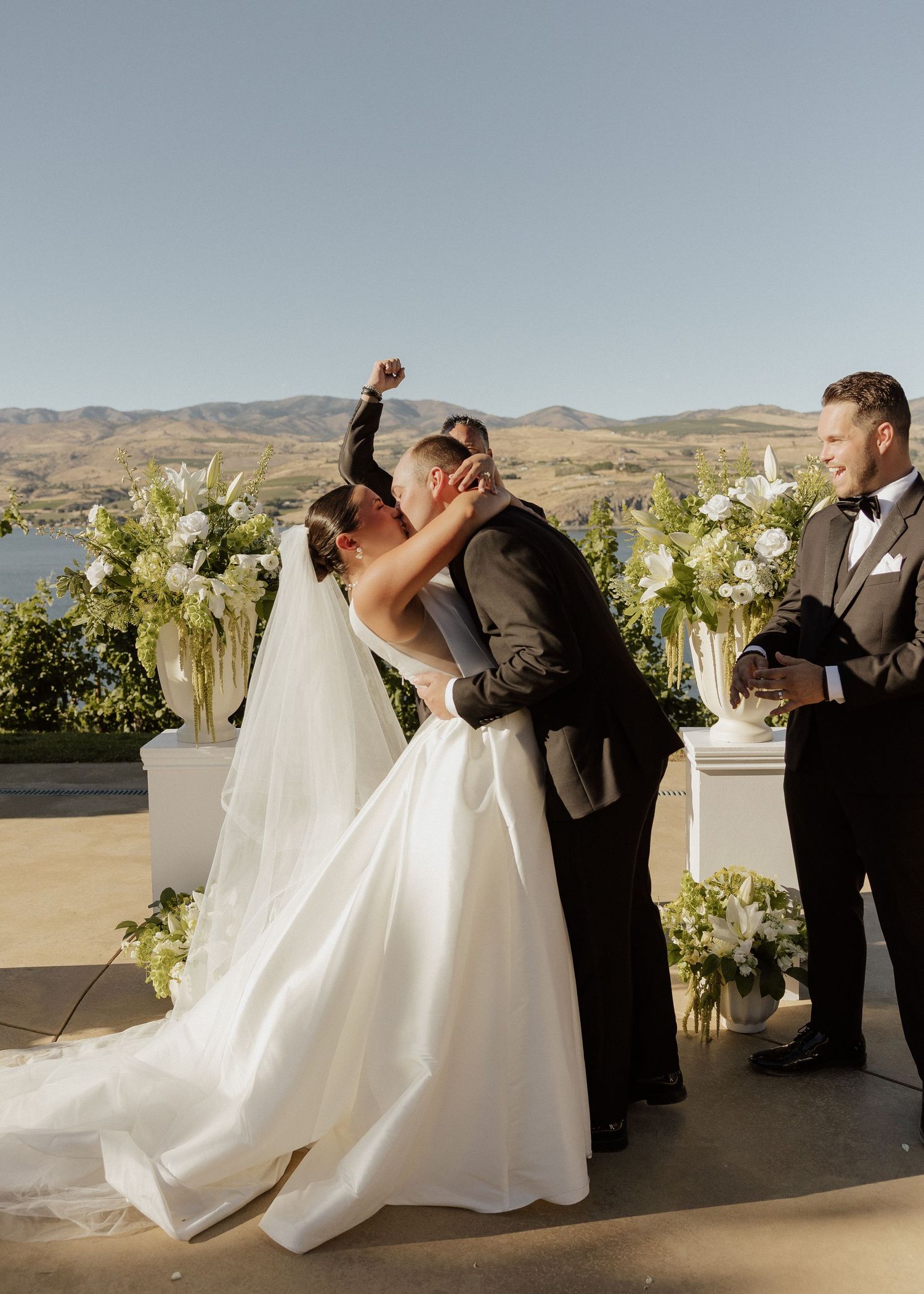 Bride and groom share first kiss at outdoor wedding ceremony with scenic lake and mountain views.
