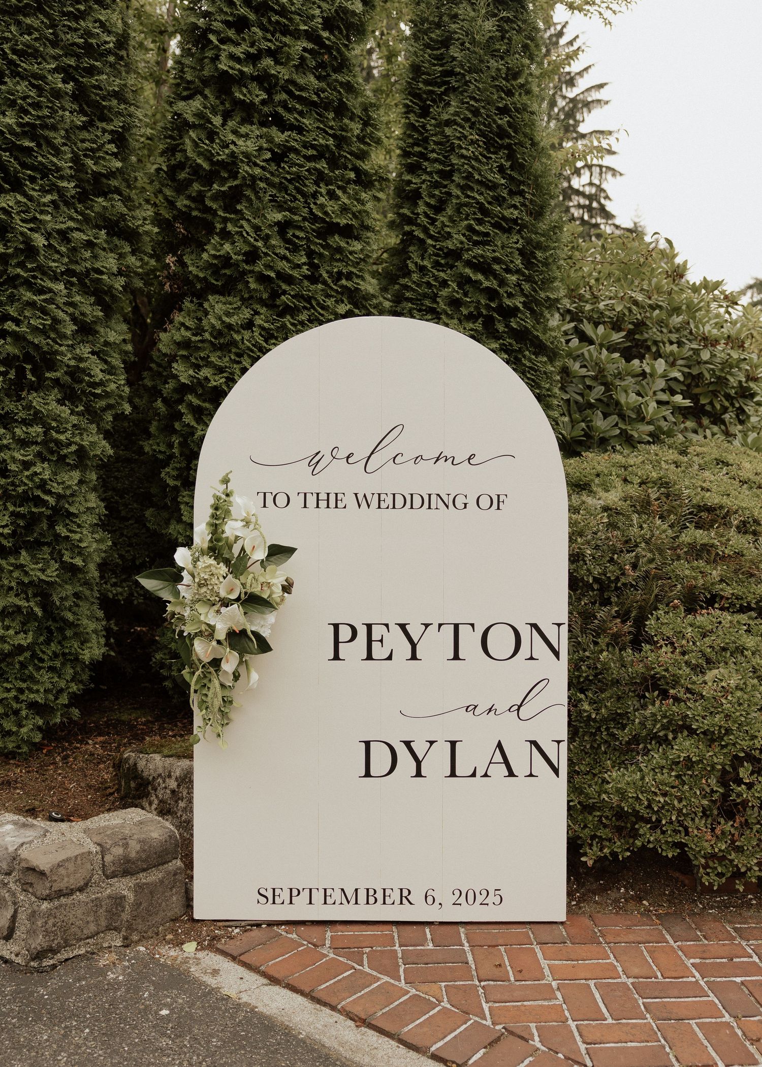 Elegant arched wedding welcome sign for Peyton and Dylan, September 6 2025, adorned with white florals against lush greenery.