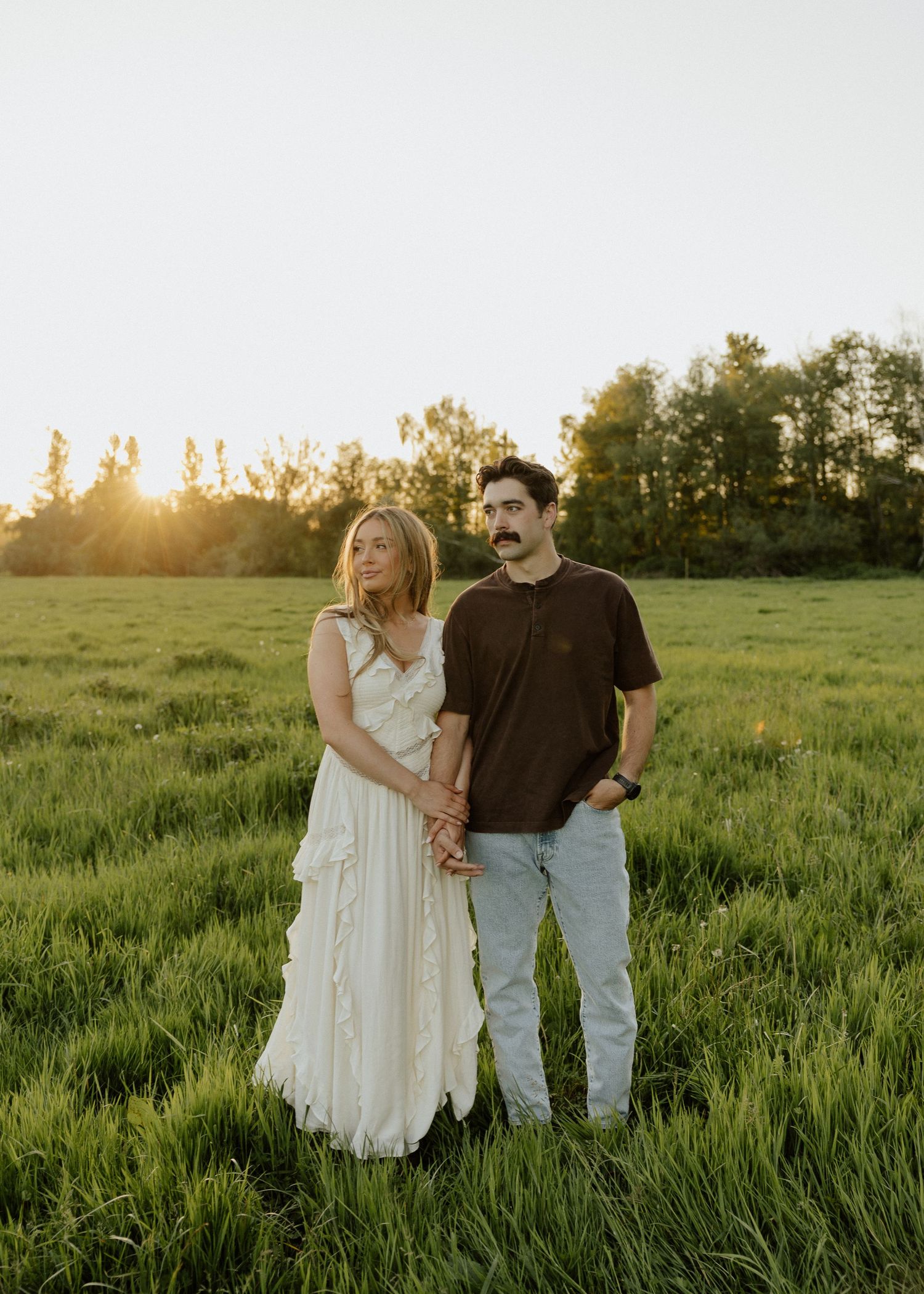 Couple standing in a lush green field at golden hour, woman in white dress, man in brown shirt and jeans.