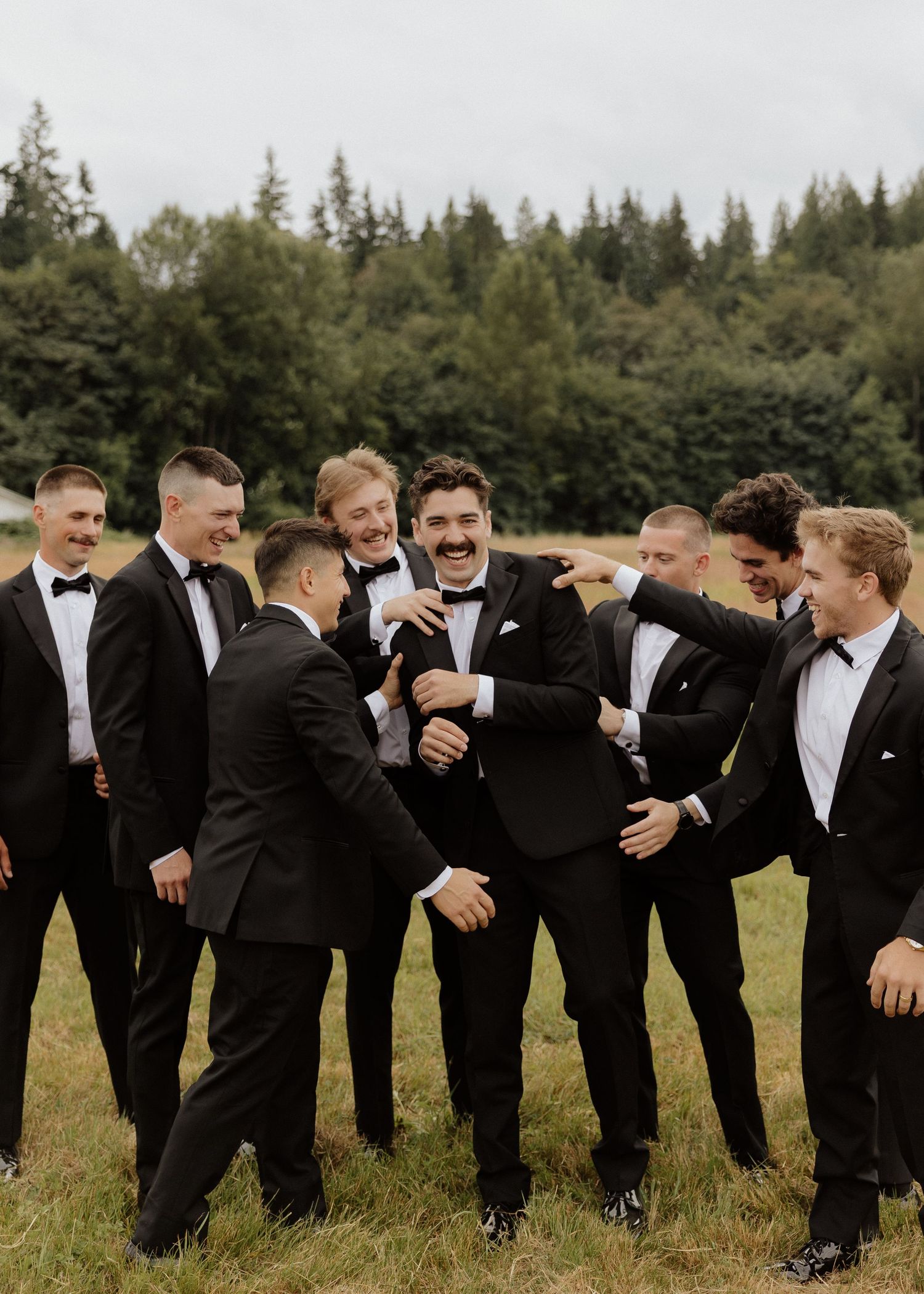 Groom and groomsmen laughing together in black tuxedos outdoors at a wedding celebration.