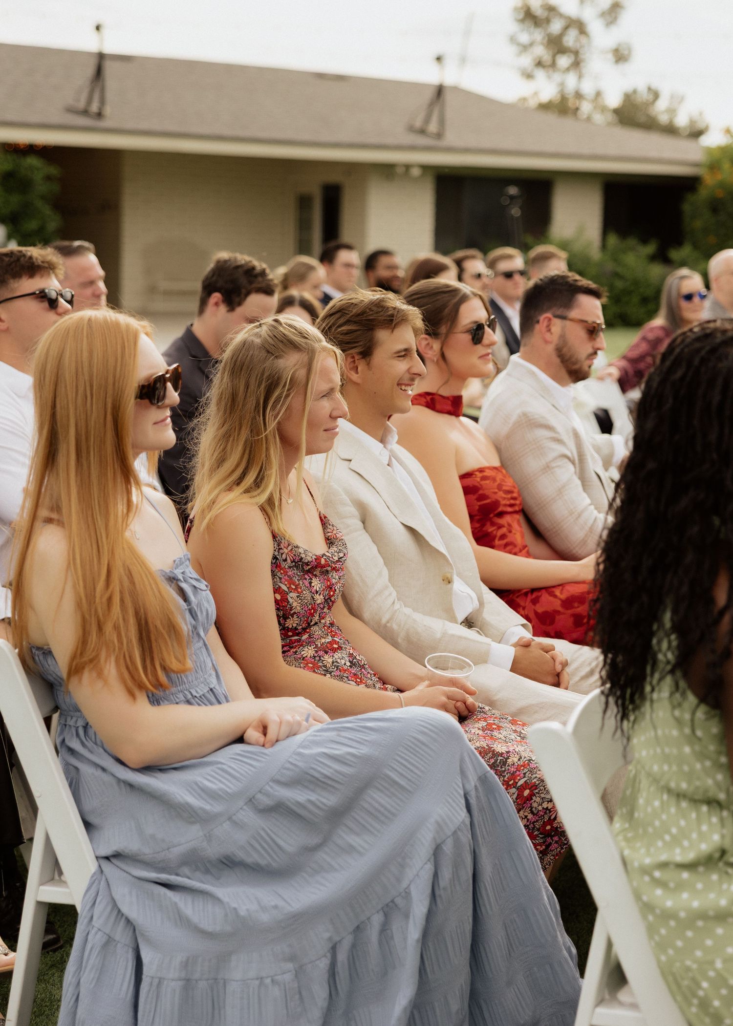 Guests seated in white chairs at an outdoor wedding ceremony, smiling and dressed in elegant summer attire.