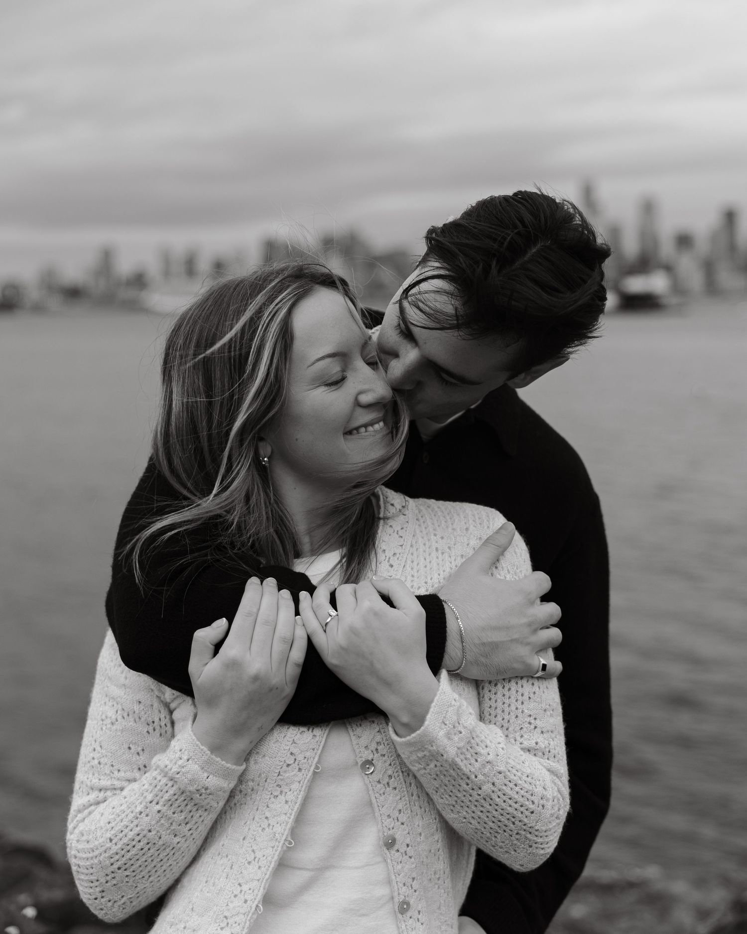 An intimate black and white moment captured of a couple embracing by the water with a cityscape behind them.