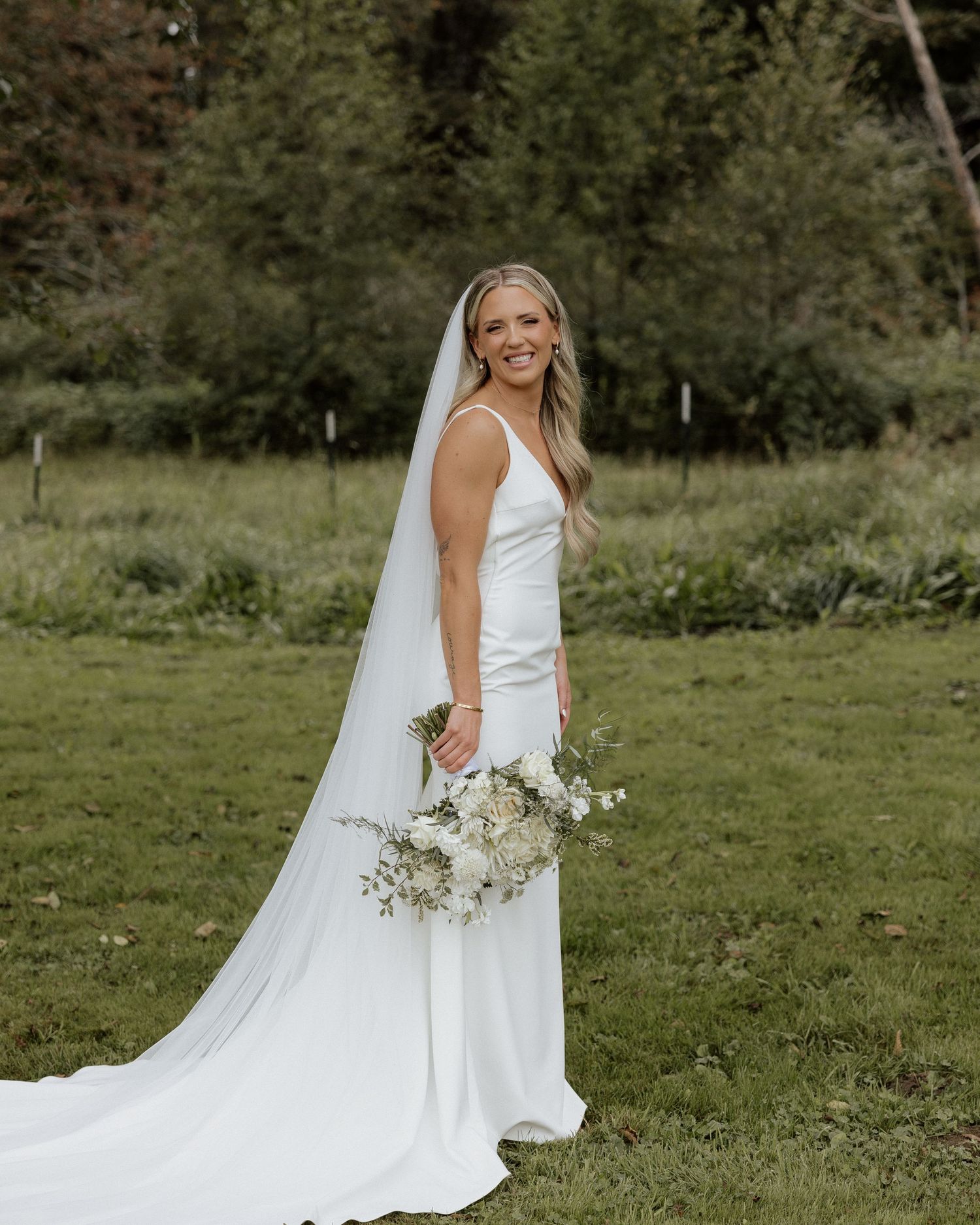 Modern bride in fitted white dress holds delicate bouquet in garden wedding portrait.
