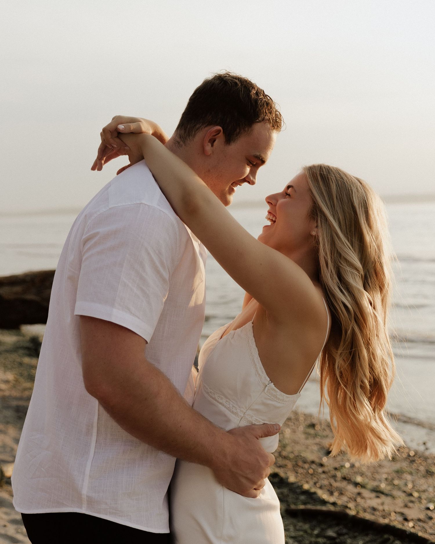 Romantic couple in white embracing on rocky beach during sunset.