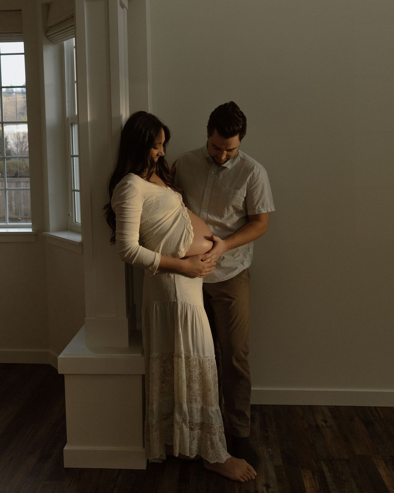 Couple sharing an intimate moment during maternity photoshoot in a rustic room with natural lighting.