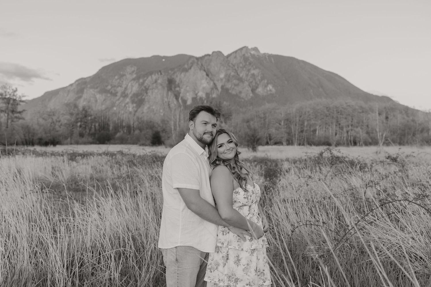 Couple embraces in grassy field with dramatic mountain landscape.