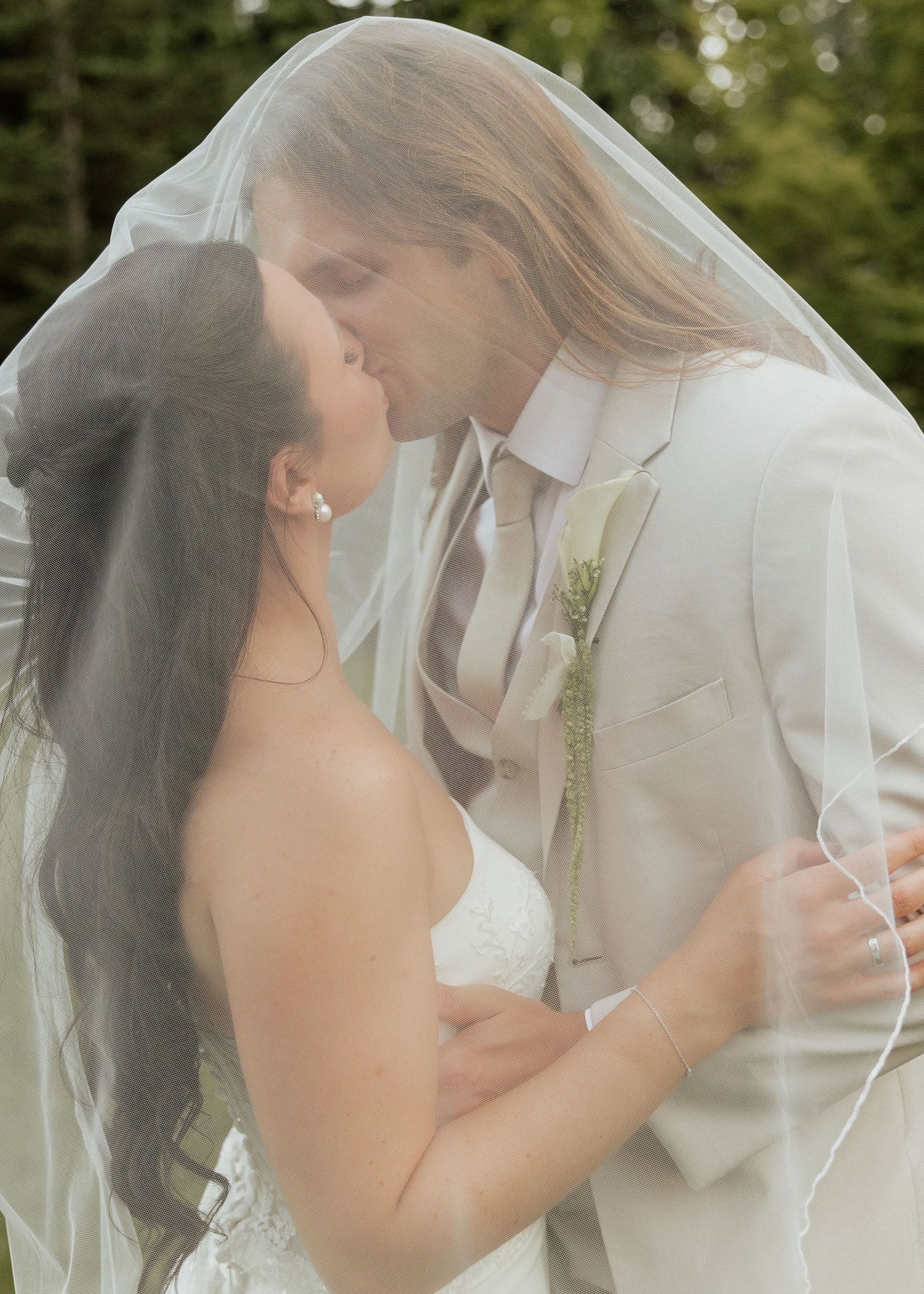Bride and groom sharing romantic kiss under flowing white veil in outdoor garden wedding portrait.