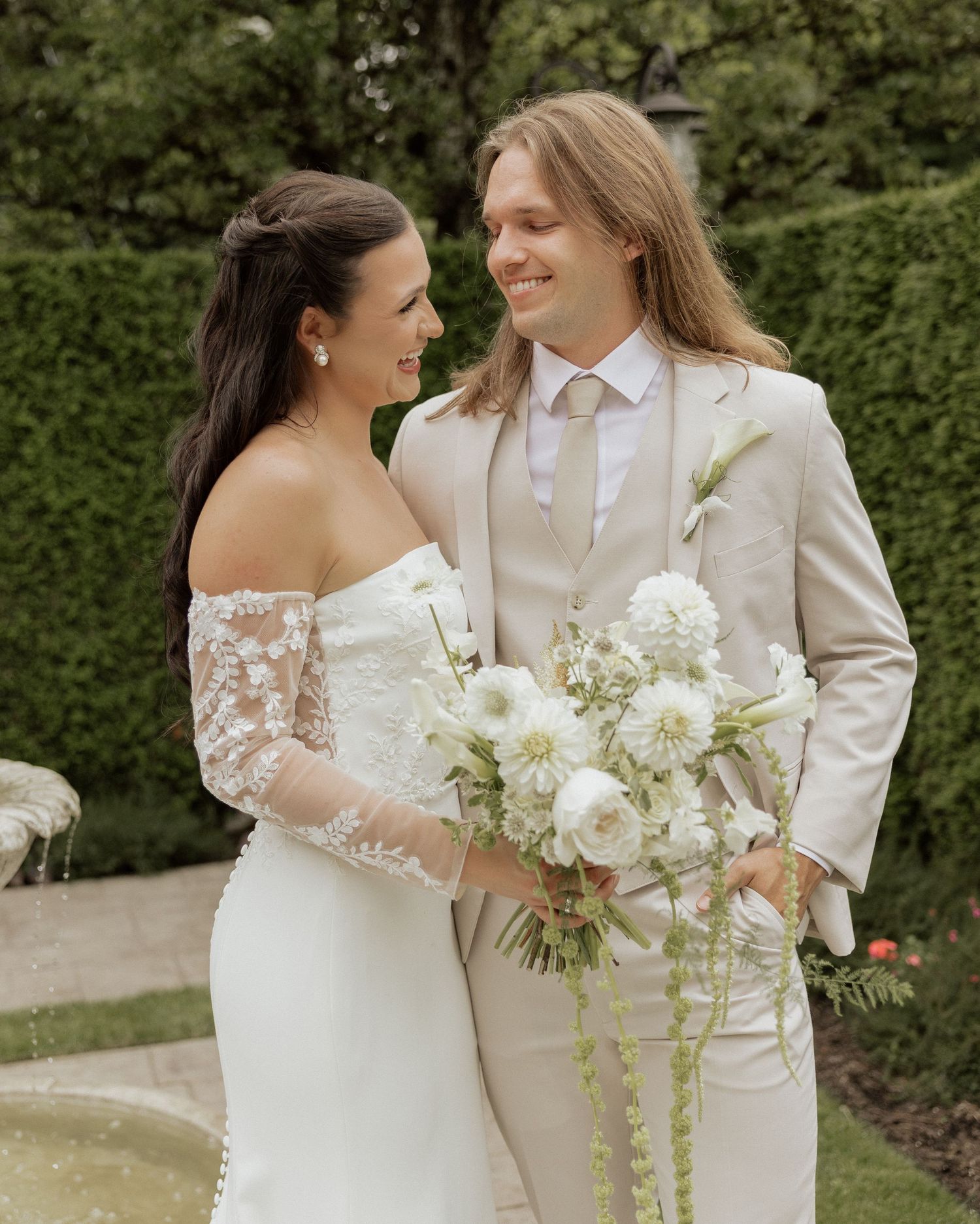 Smiling bride and groom in beige suit sharing romantic moment while holding white floral bouquet in garden setting.