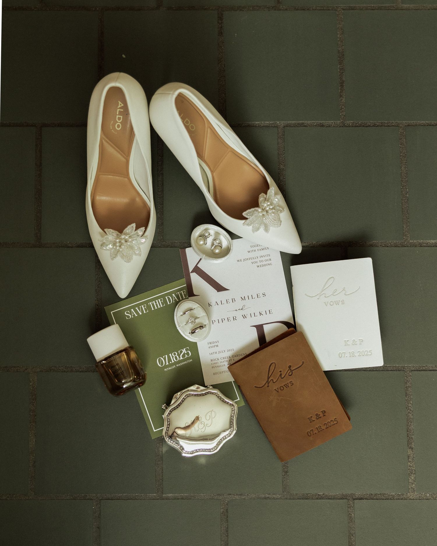 White bridal flats with bow detail displayed with wedding stationery, perfume, and accessories on green tile.