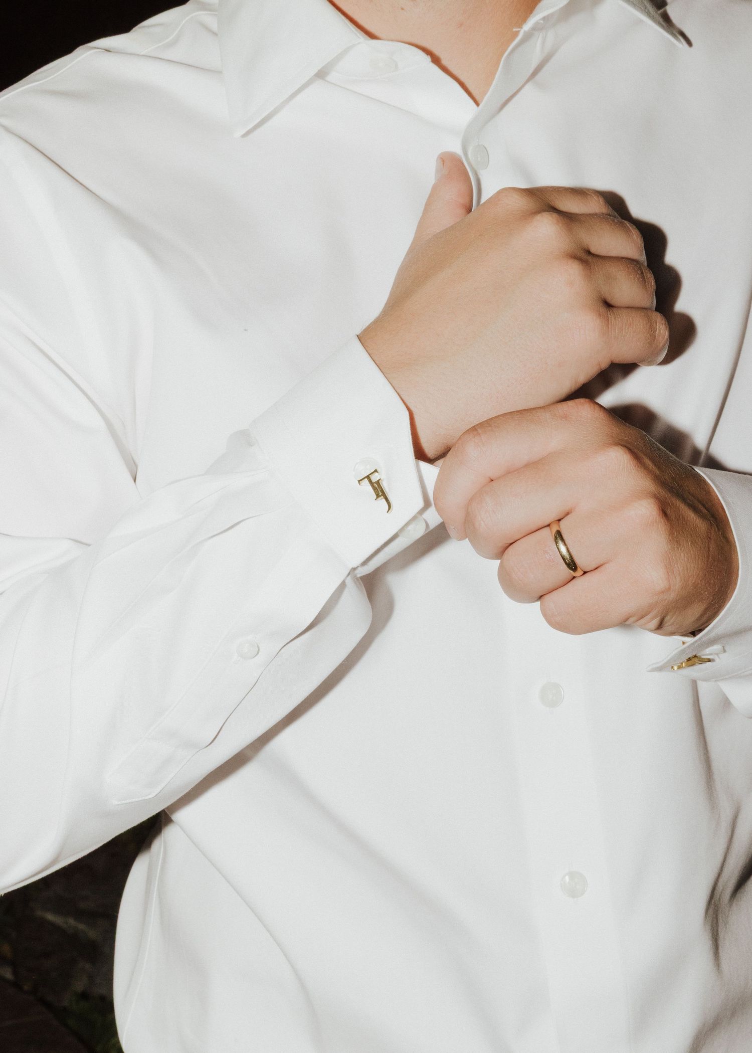 Groom in white dress shirt fastening gold cufflinks, wearing a gold wedding band on his ring finger.