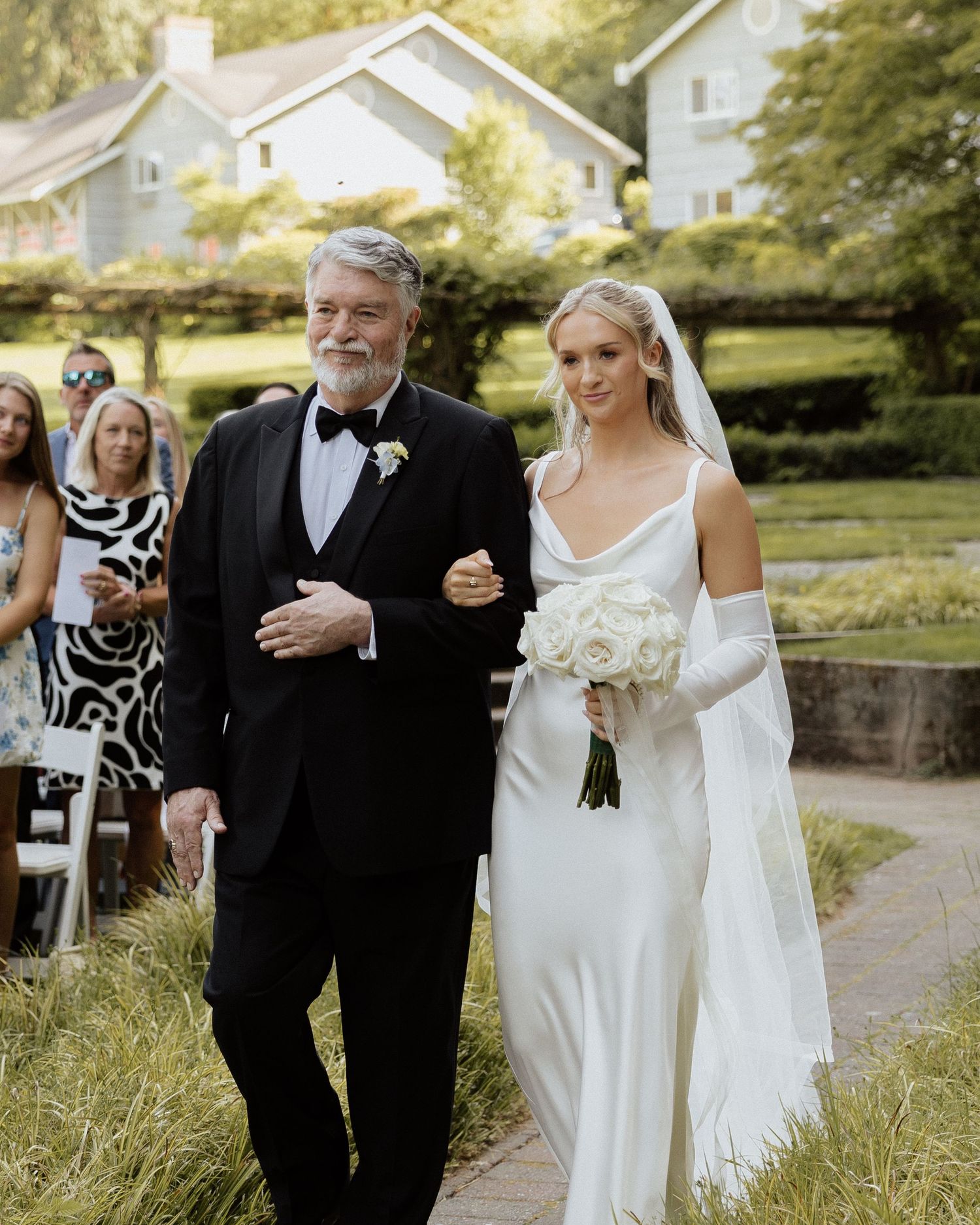 Bride in white satin gown walks down aisle with father in tuxedo at outdoor wedding ceremony.