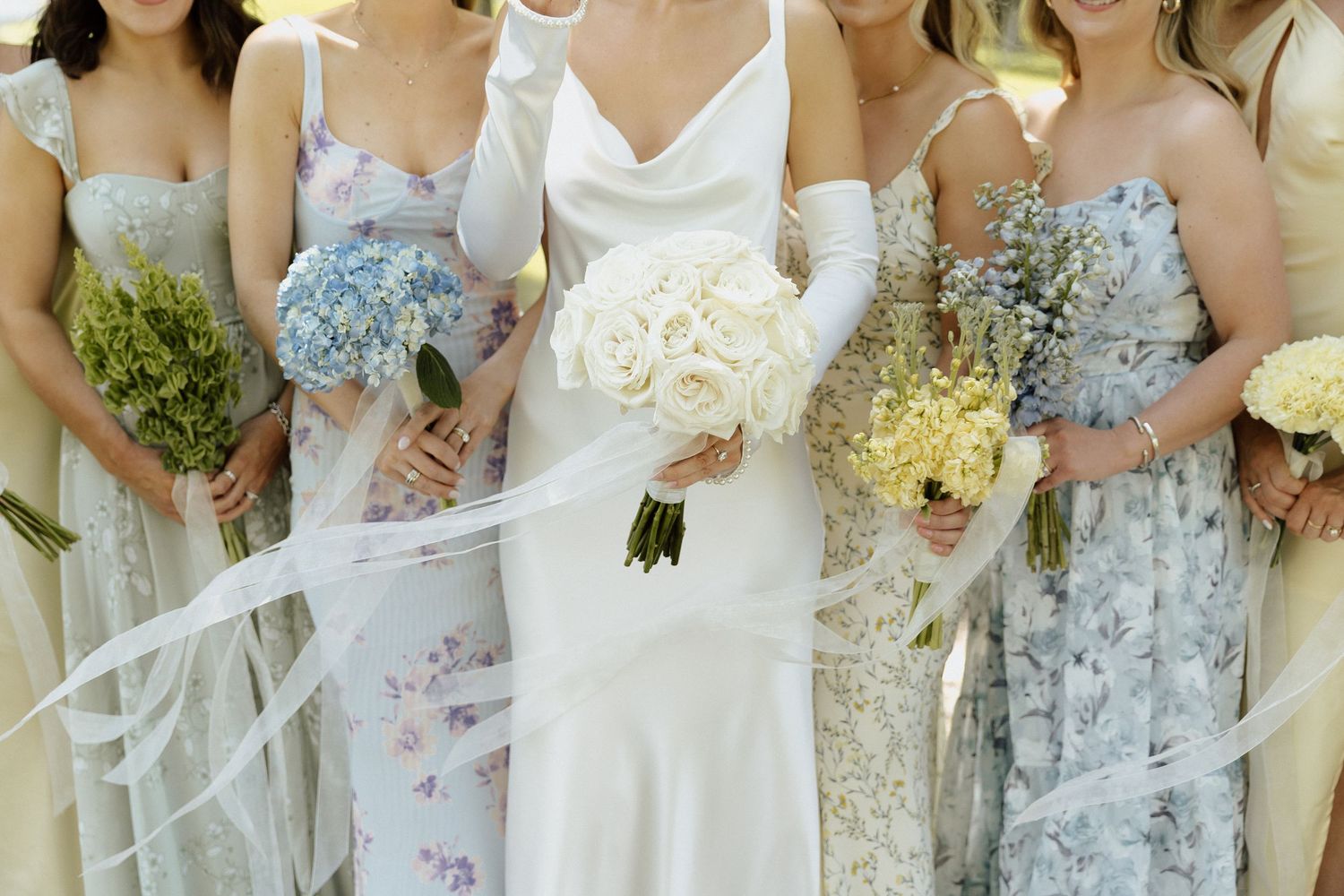 Bride in white gown holding rose bouquet with bridesmaids in floral dresses holding colorful flower bouquets outdoors.