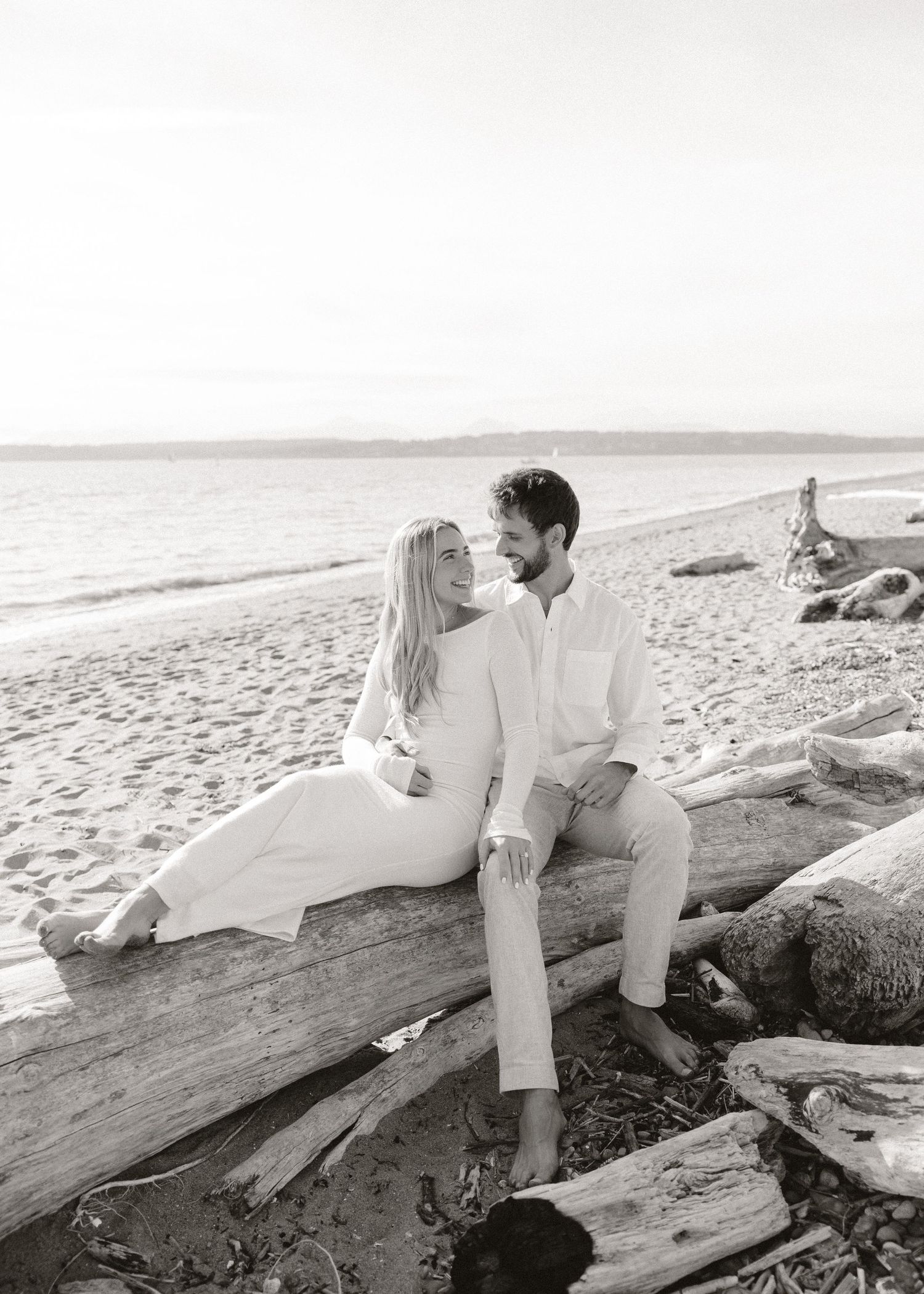 Black and white photo of a couple sitting on driftwood on a beach, smiling at each other in white outfits.