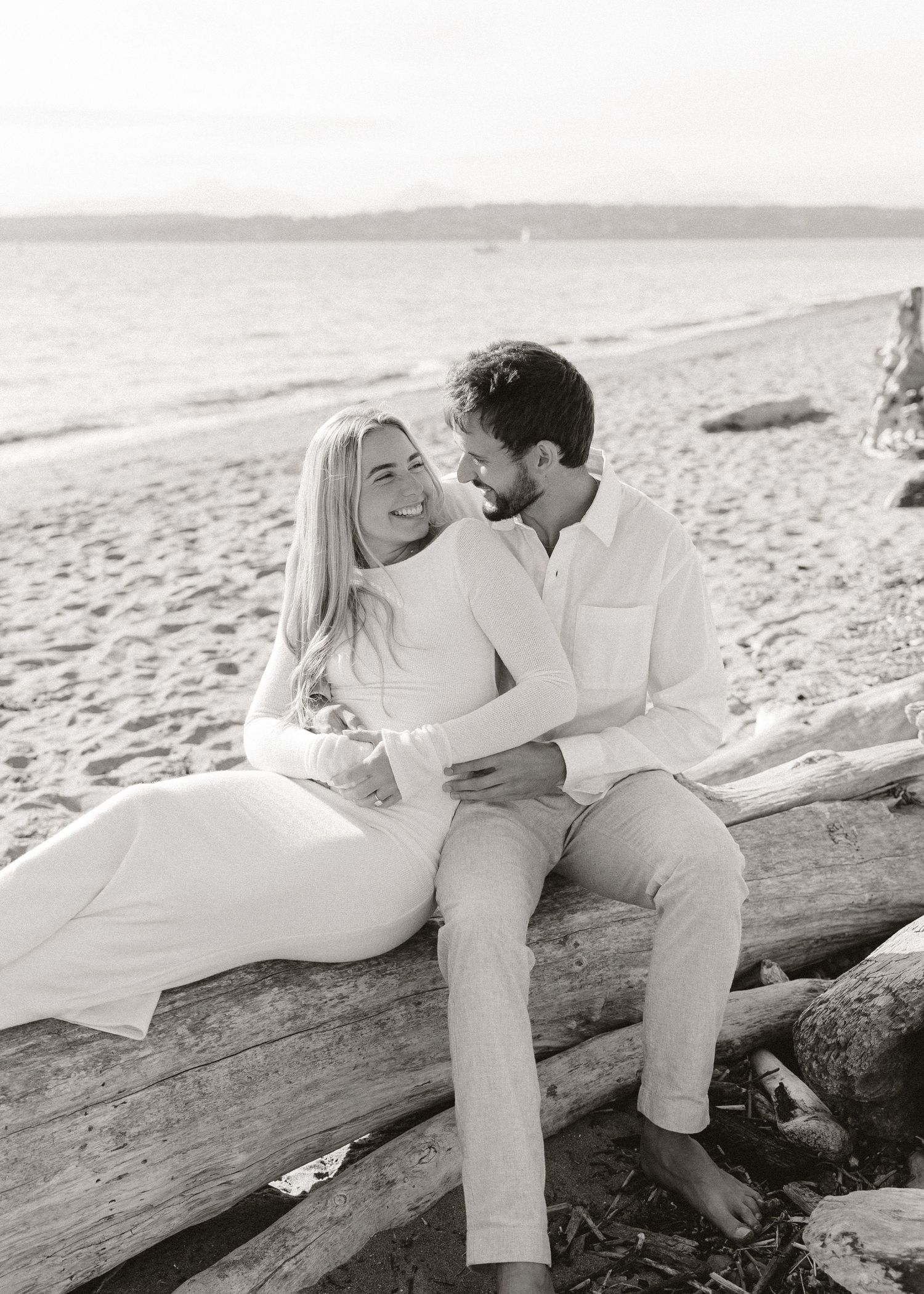 Black and white photo of a couple sitting on driftwood at the beach, smiling and embracing romantically.