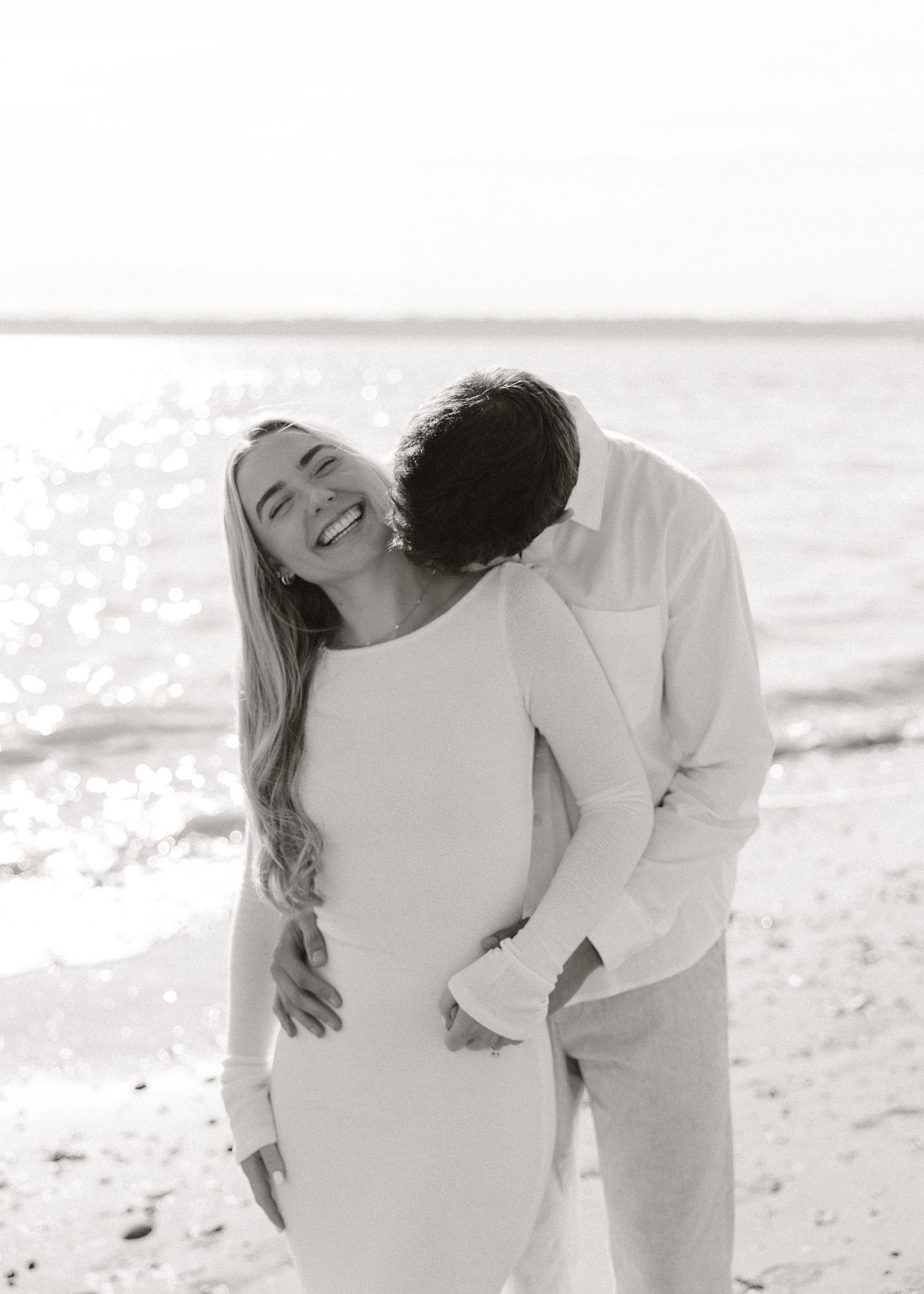Couple embracing on beach, woman laughing joyfully as man kisses her neck, black and white photo.