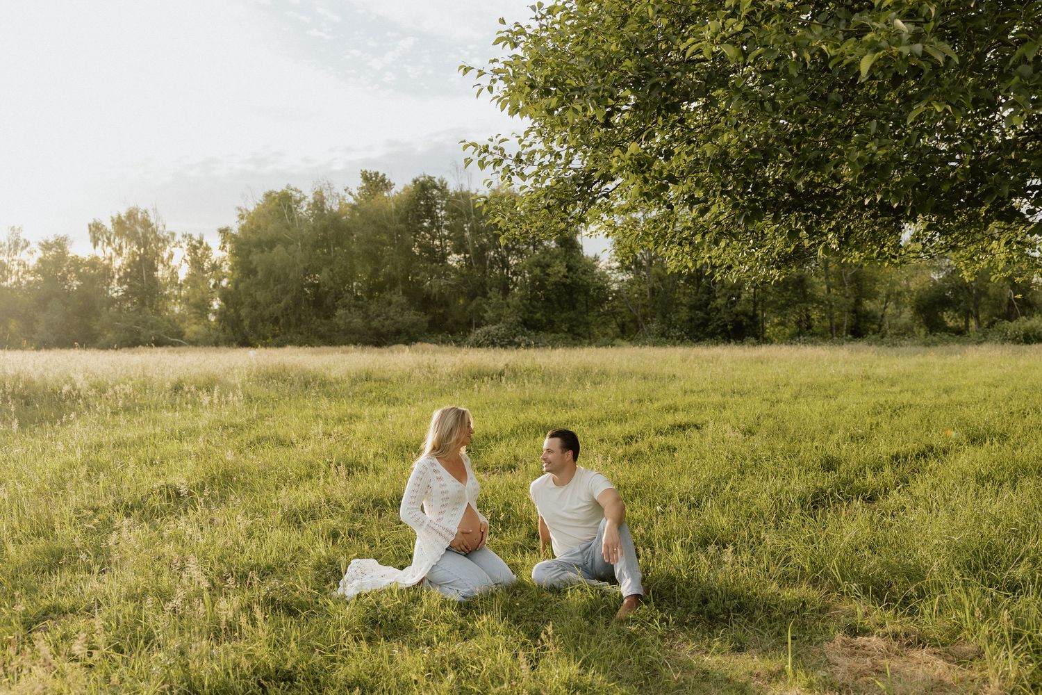 Couple sitting on green grass in a sunny field, wearing white outfits, facing each other near a large tree.