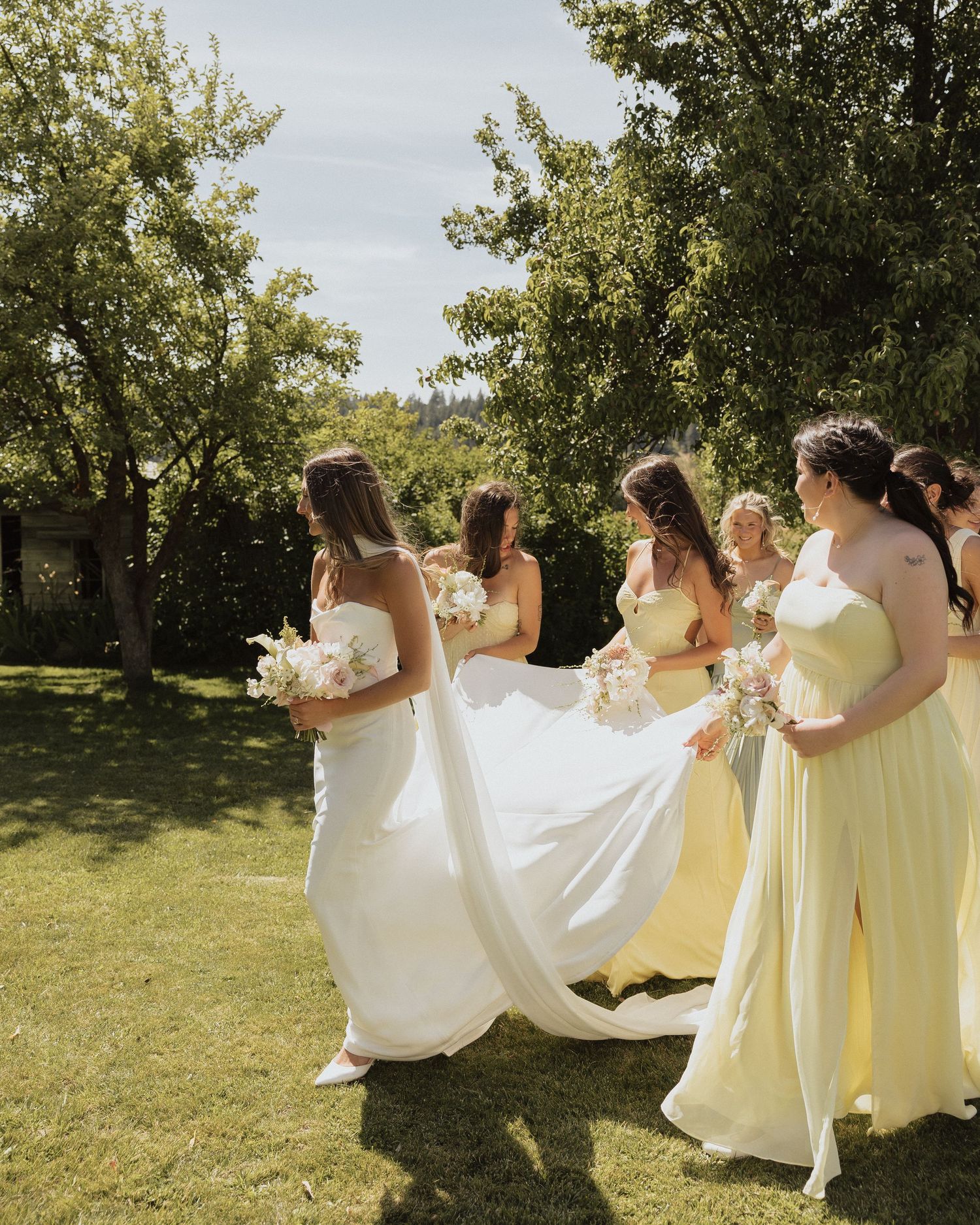 Bride in white gown walks with bridesmaids in yellow dresses carrying bouquets outdoors on sunny day.