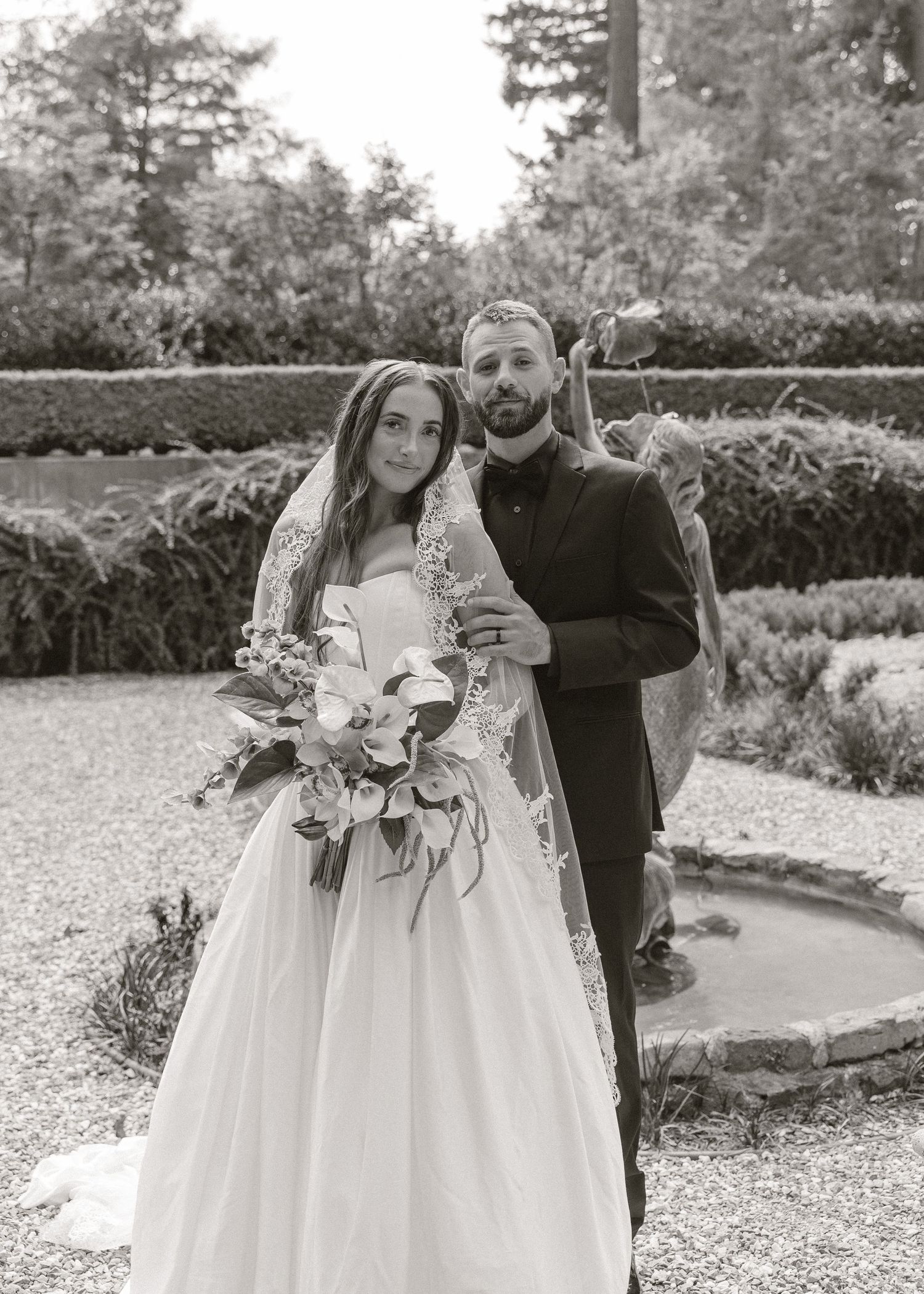 Black and white wedding photo of bride holding bouquet and groom in suit posing in elegant garden.